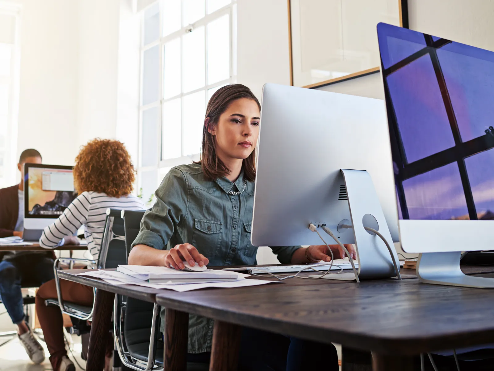 Young professionals working intently on desktop computers in a bright modern office space with large windows.
