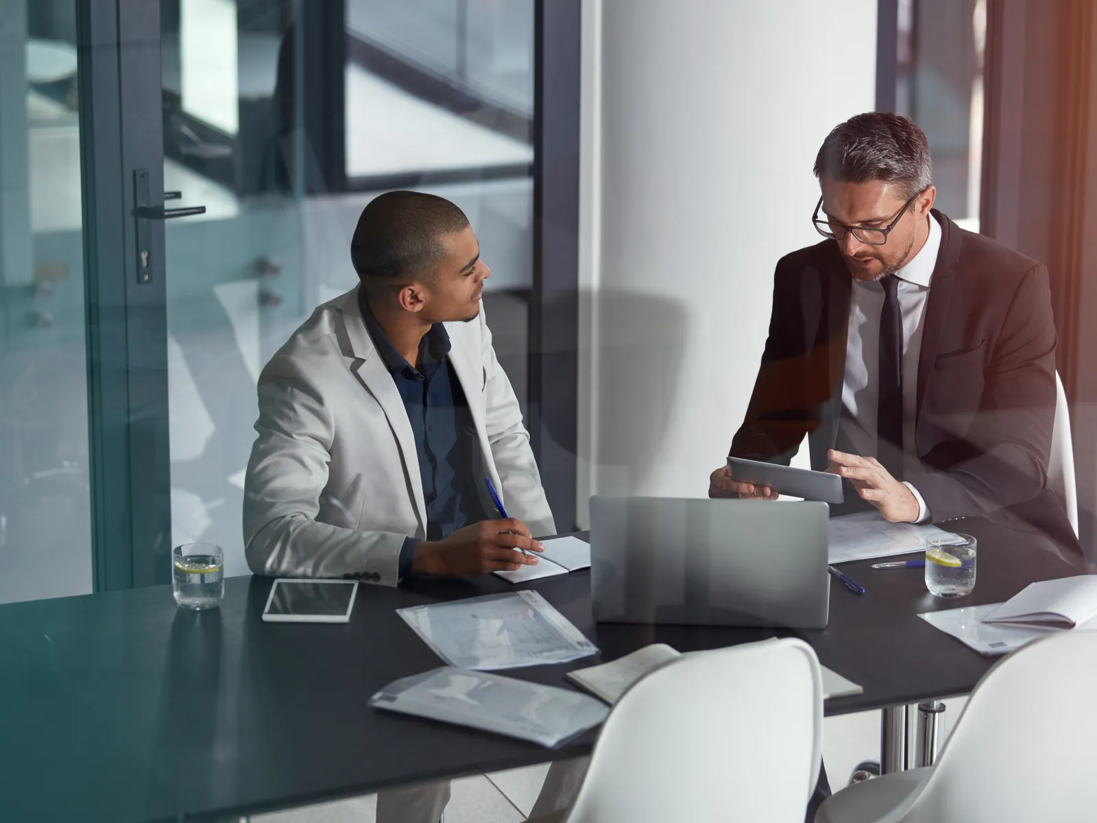 Two businessmen in suits discussing documents and tablet at office meeting table with laptop and papers