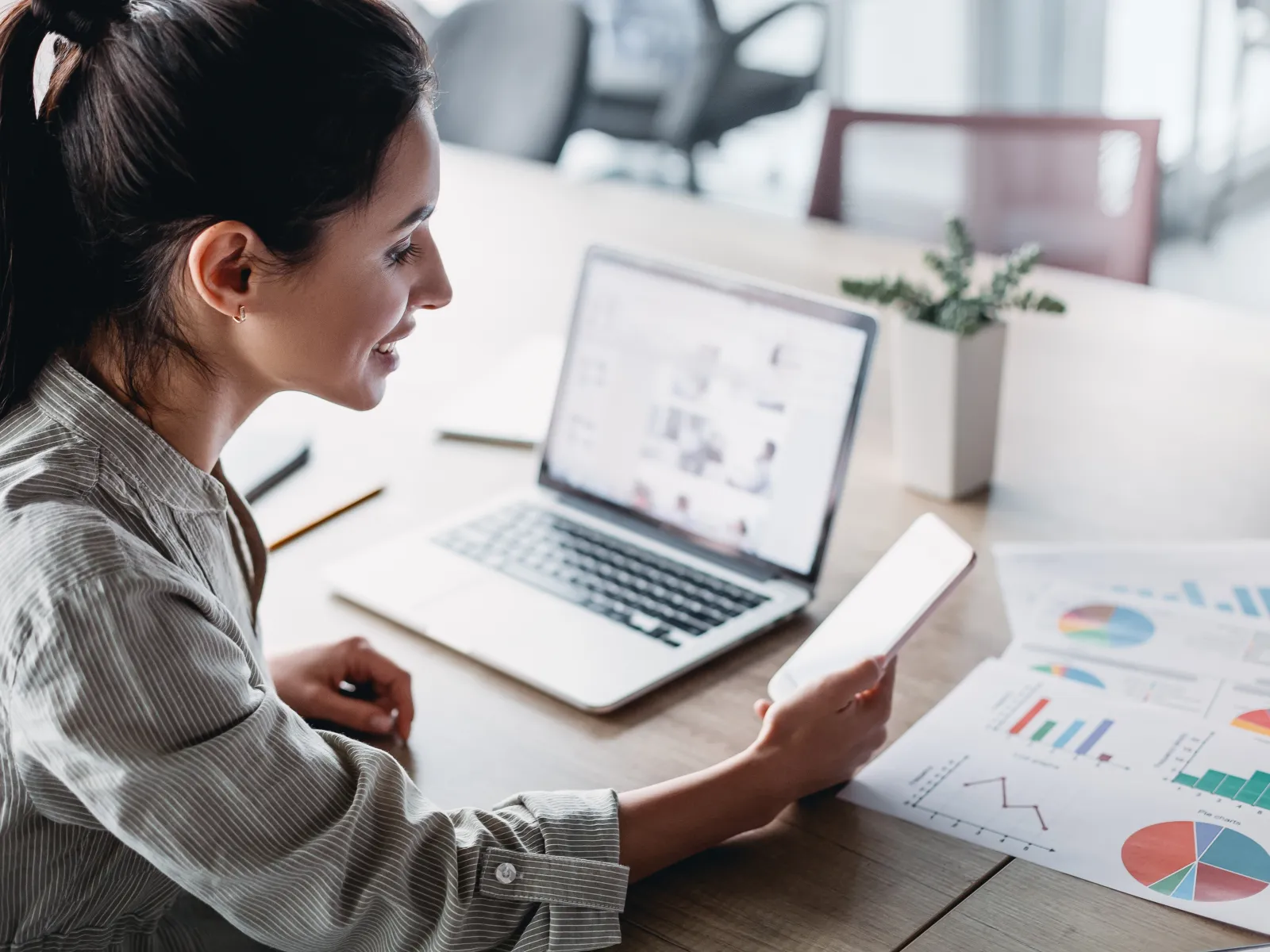 Young woman using smartphone and laptop analyzing financial charts and graphs at a light office desk.