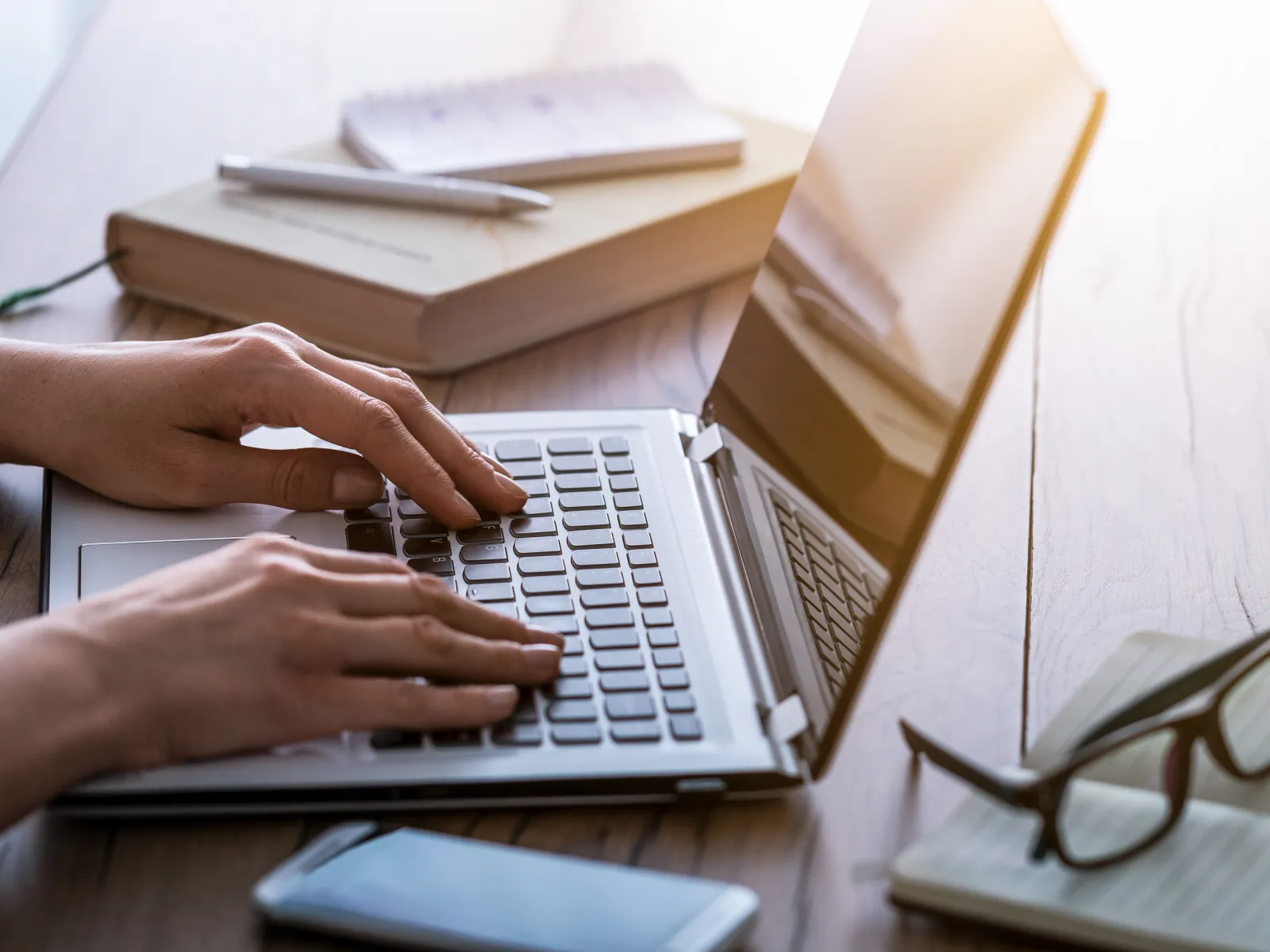 Hands typing on a laptop keyboard on a wooden desk with glasses, notebook, smartphone, and books nearby.