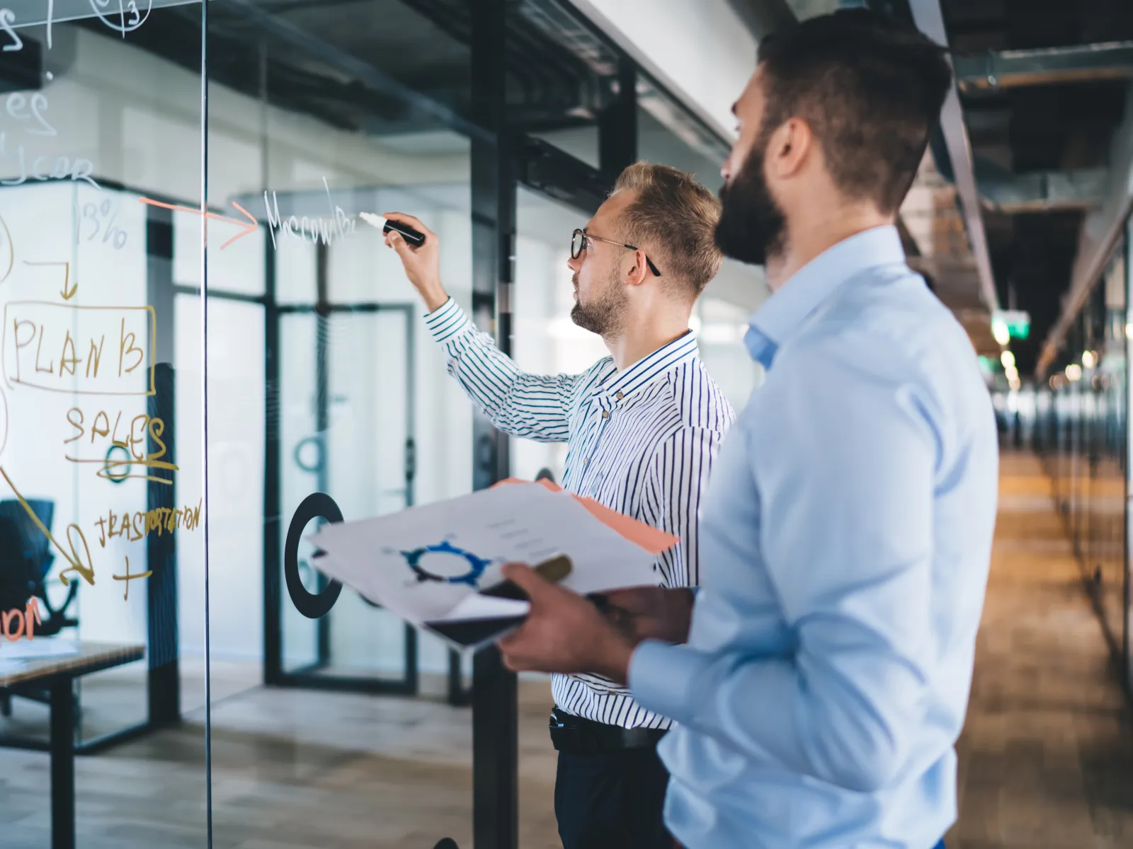 Two business professionals brainstorming and writing plans on a glass wall in a modern office.