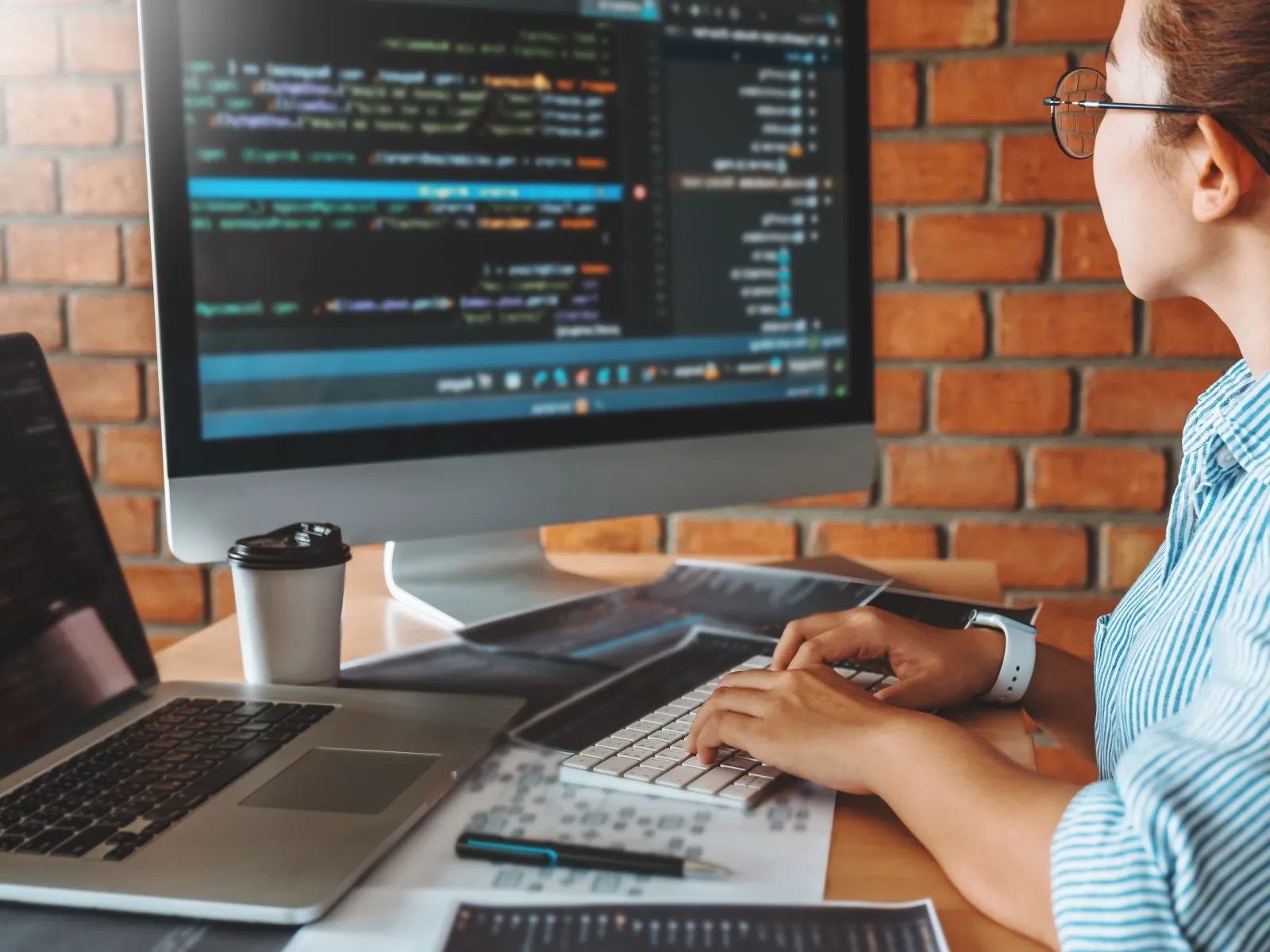 Person coding on a desktop computer with a laptop and coffee cup on a wooden desk in a brick-walled room.