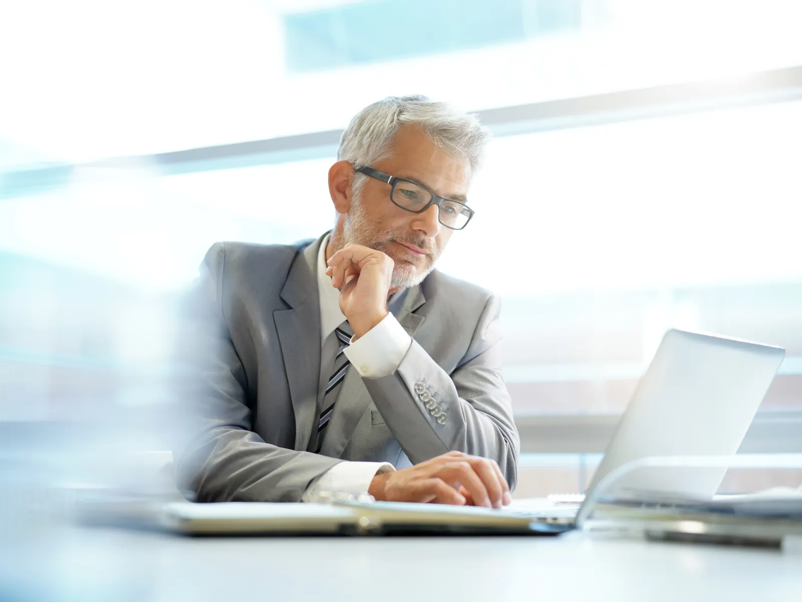 Mature businessman in gray suit working thoughtfully on laptop in bright modern office.