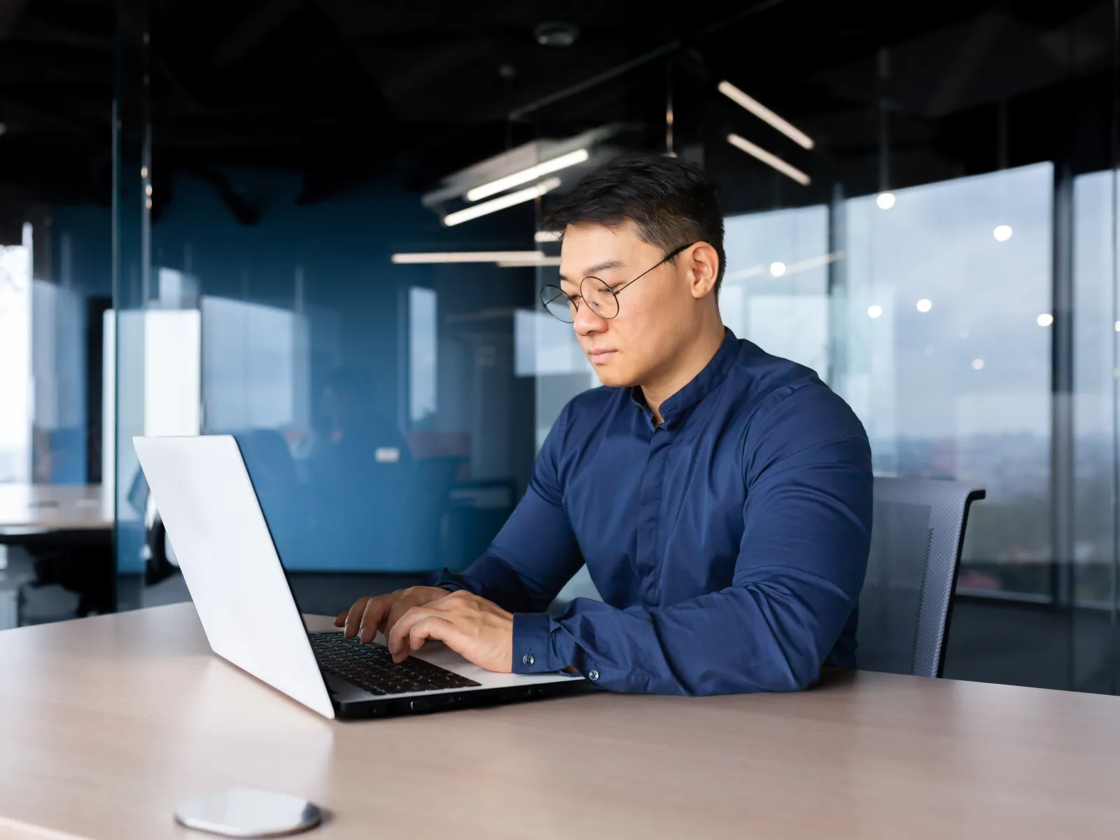 Focused man in blue shirt working on laptop at modern office desk with glass walls and city view.