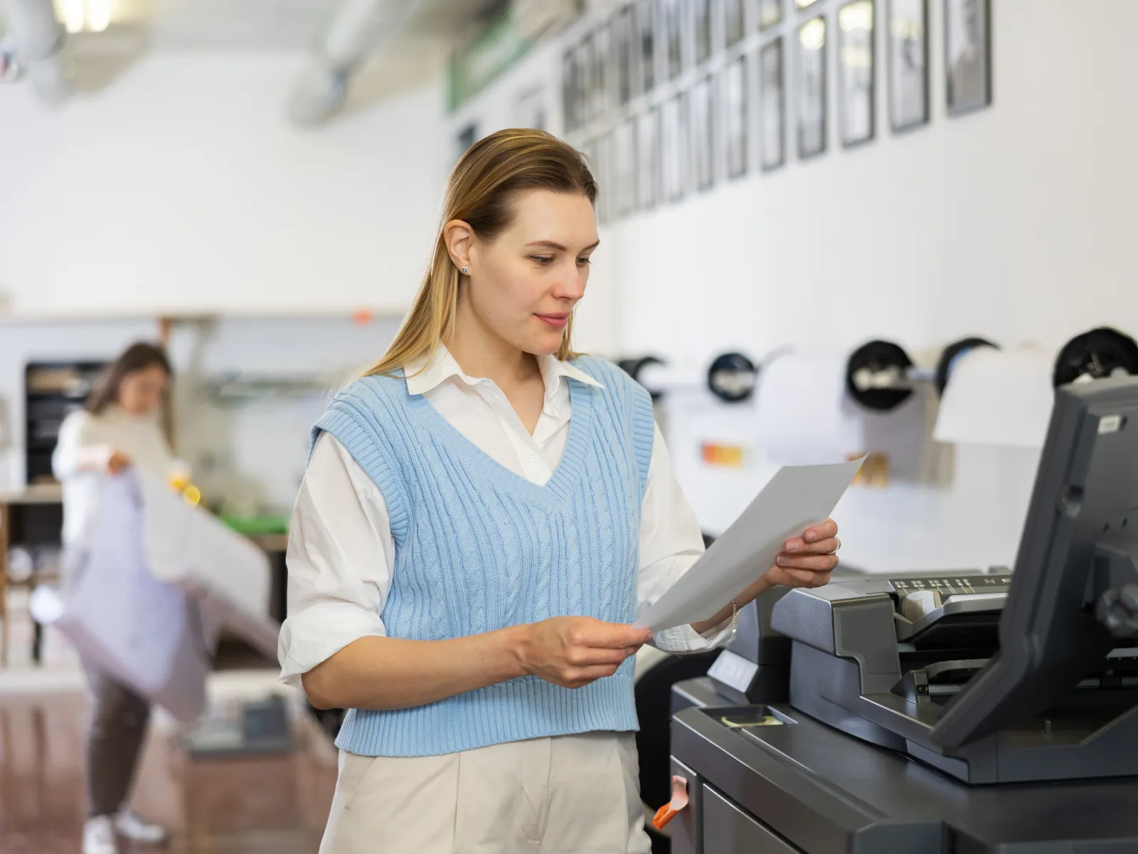 Woman in blue vest checking printed paper next to a photocopier in a bright office space.