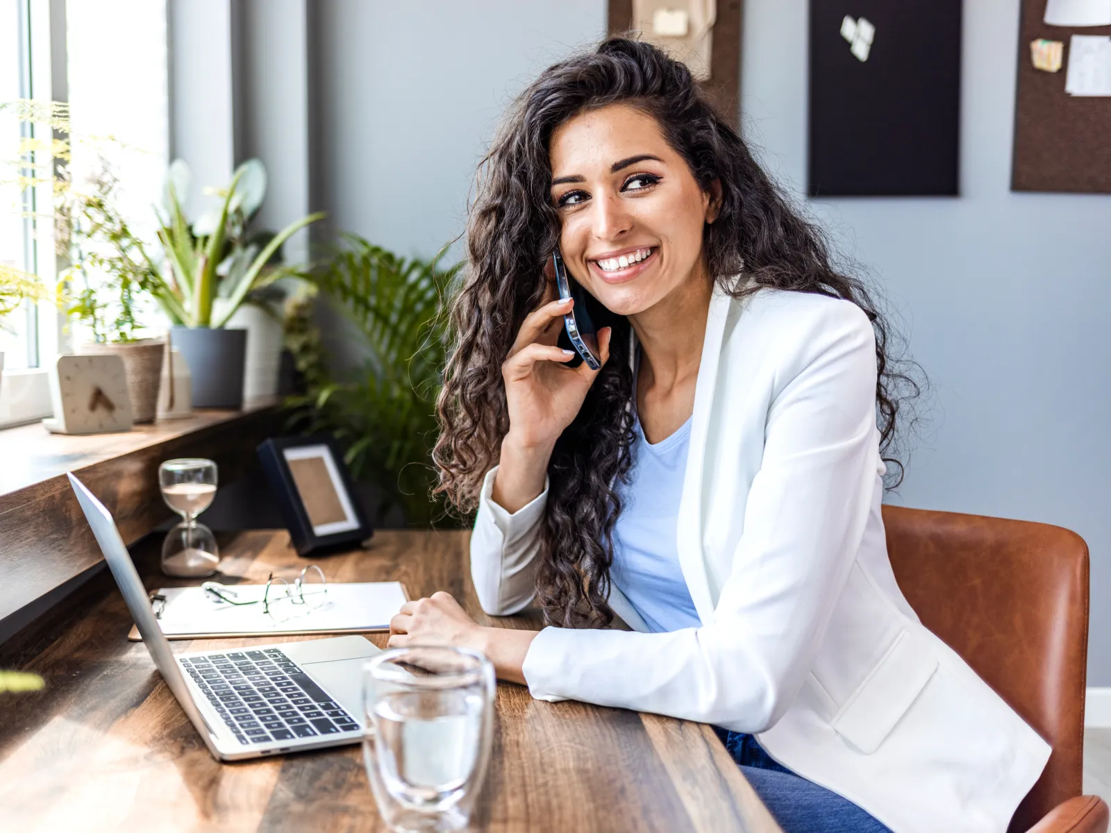 Smiling woman in white blazer talks on phone while working on laptop at wooden desk with plants nearby