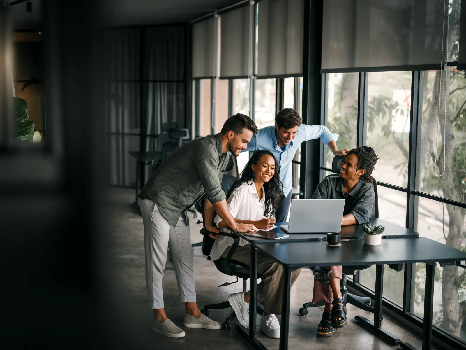 Four diverse coworkers collaborating happily around a laptop in a modern office with large windows.