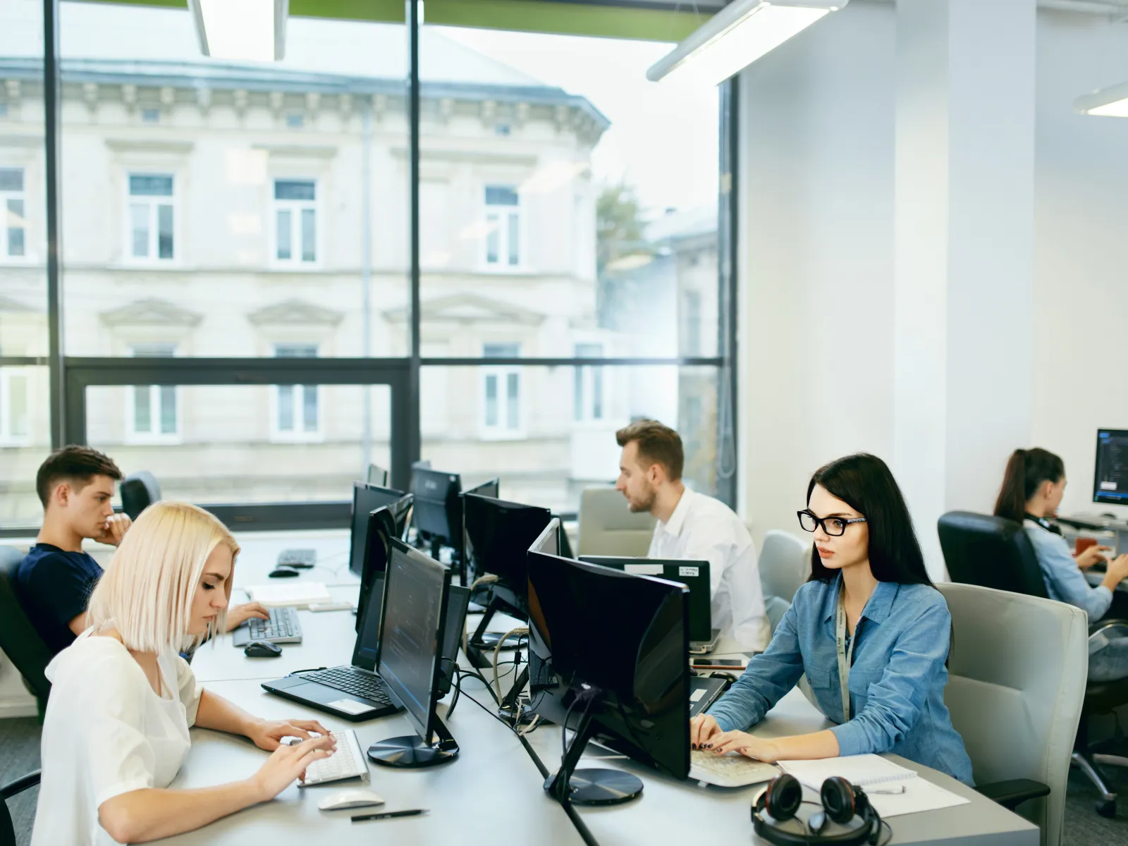 Young professionals working at computers in a modern office with large windows and natural light.