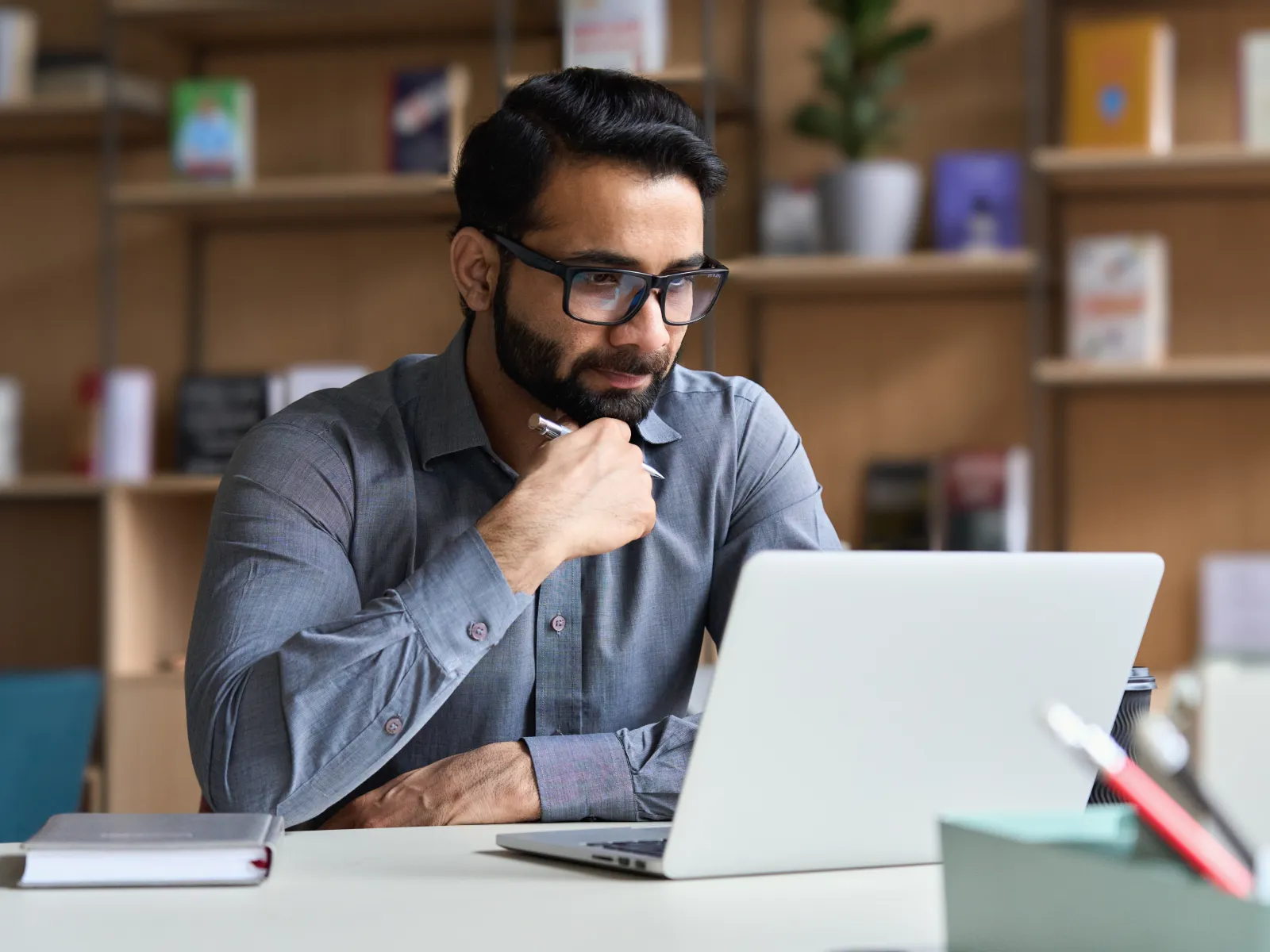 Man wearing glasses working thoughtfully on a laptop in a modern office setting with bookshelves in the background