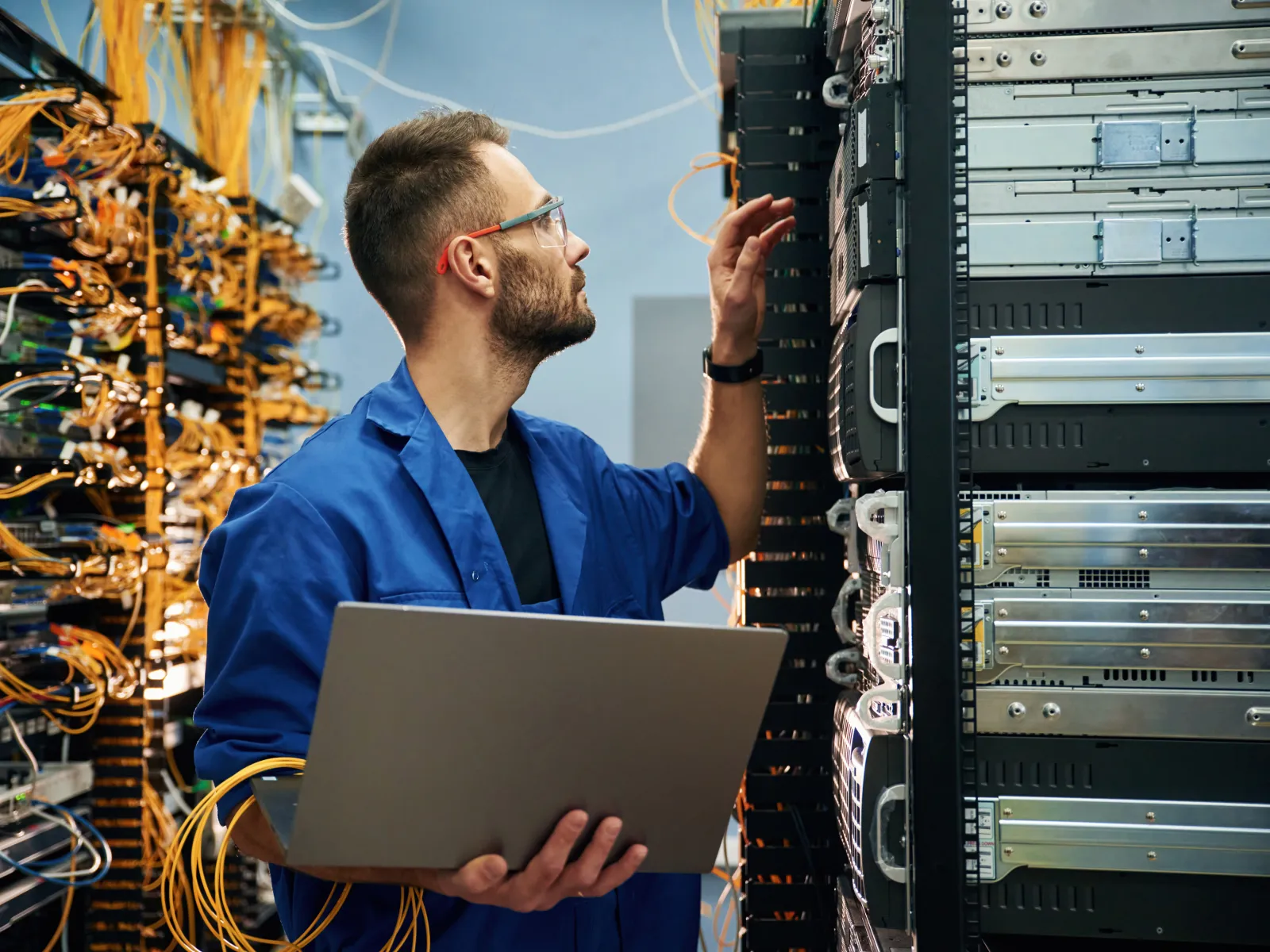 Technician in blue coat holding laptop while checking server hardware in data center with network cables.