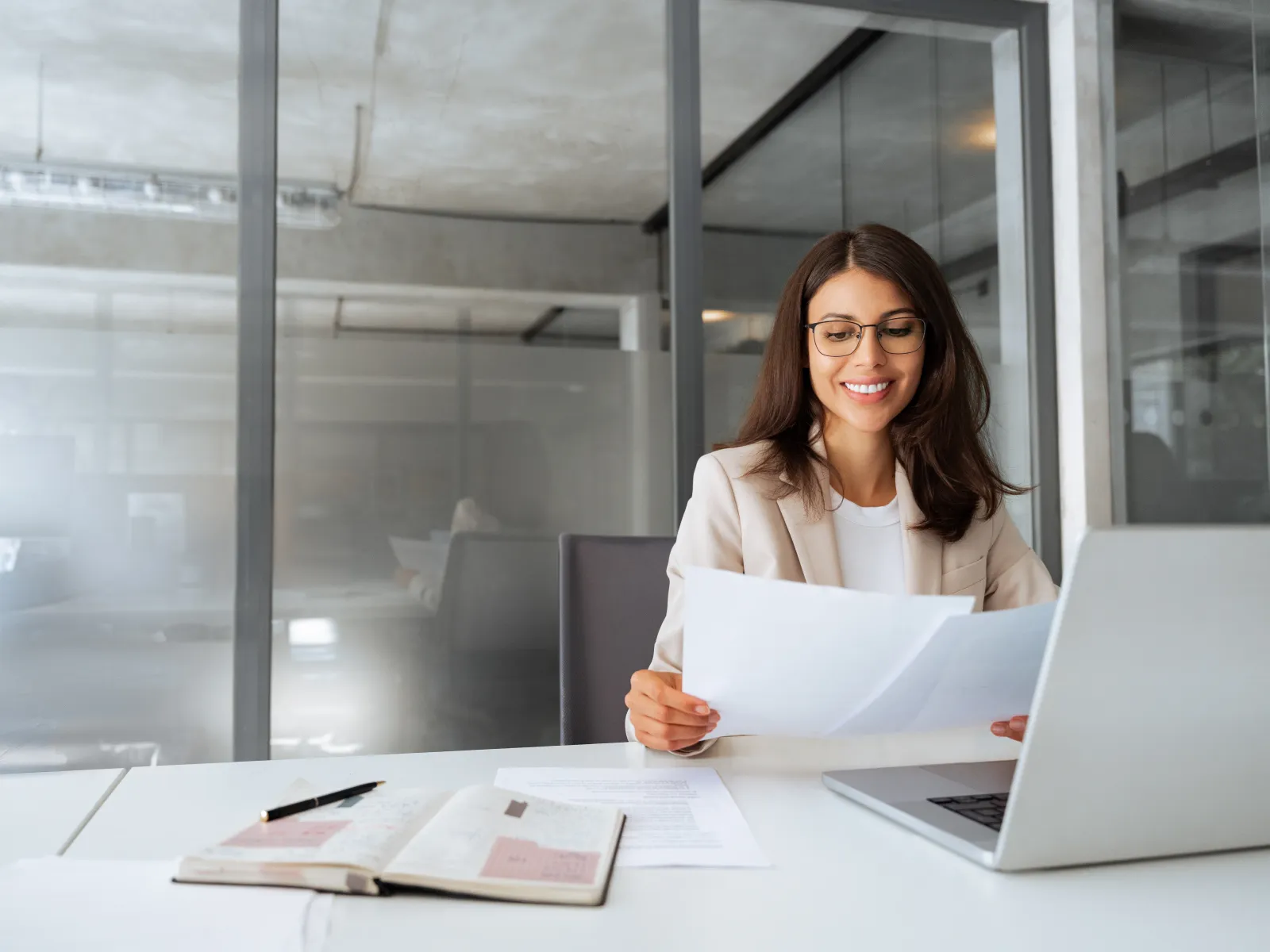 Smiling woman in glasses and blazer reviewing documents at desk with laptop and open notebook in modern office.