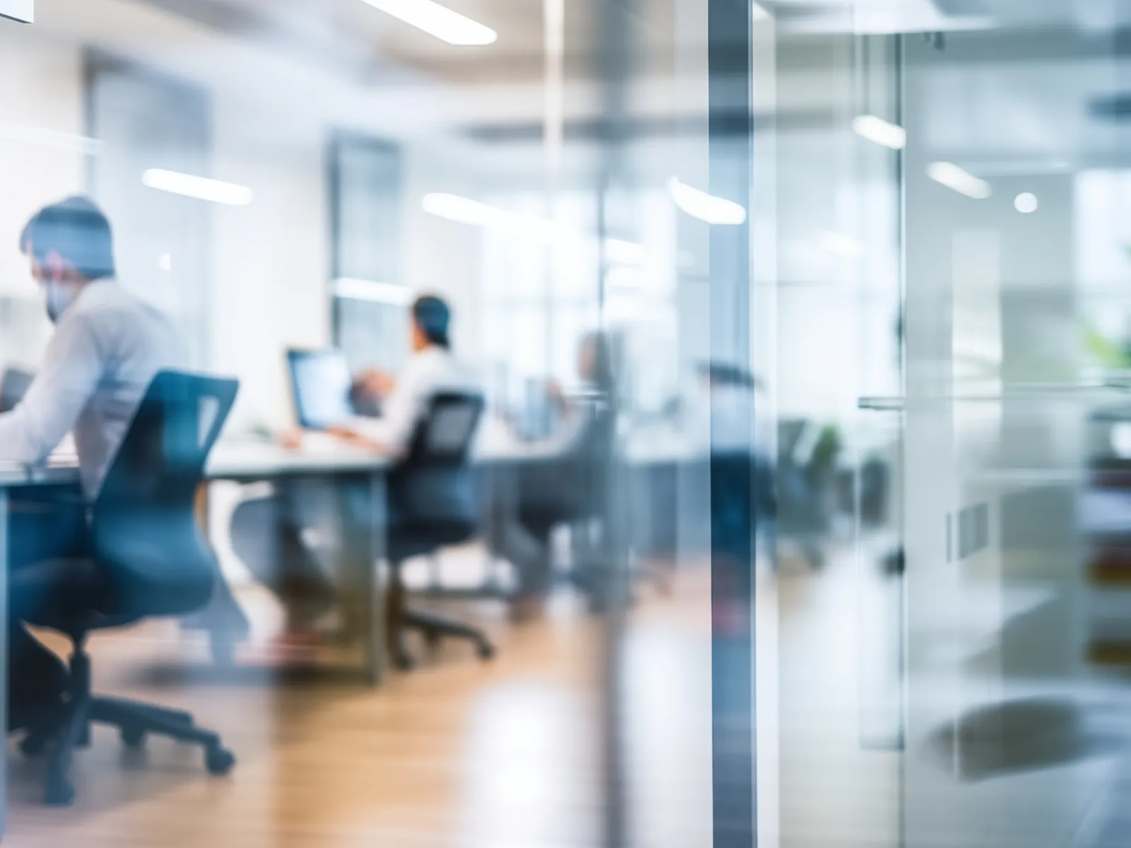Blurred view of professionals working on laptops in a modern glass-walled office environment.