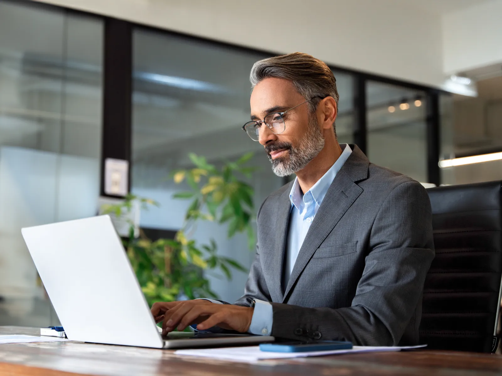 Middle-aged businessman in suit working on laptop in modern office with plants and glass walls.