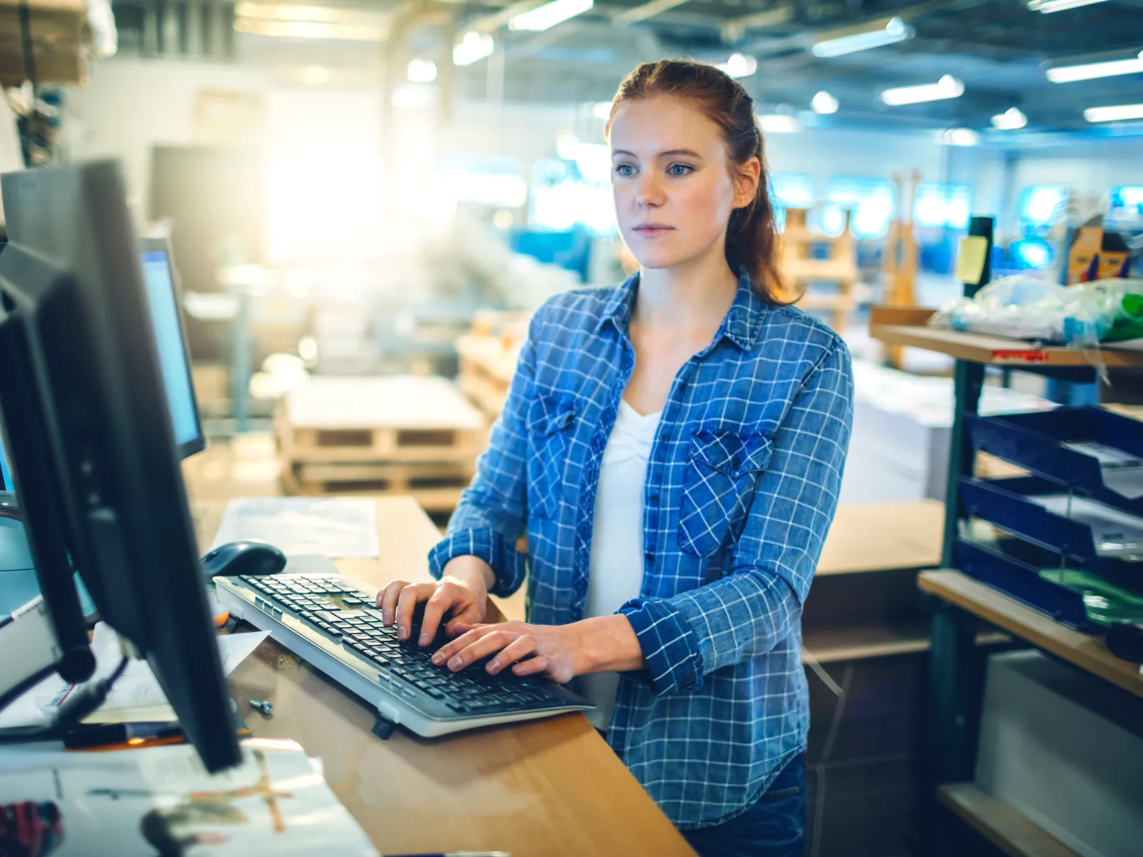 Young woman working on computer at standing desk in industrial warehouse or workshop environment.