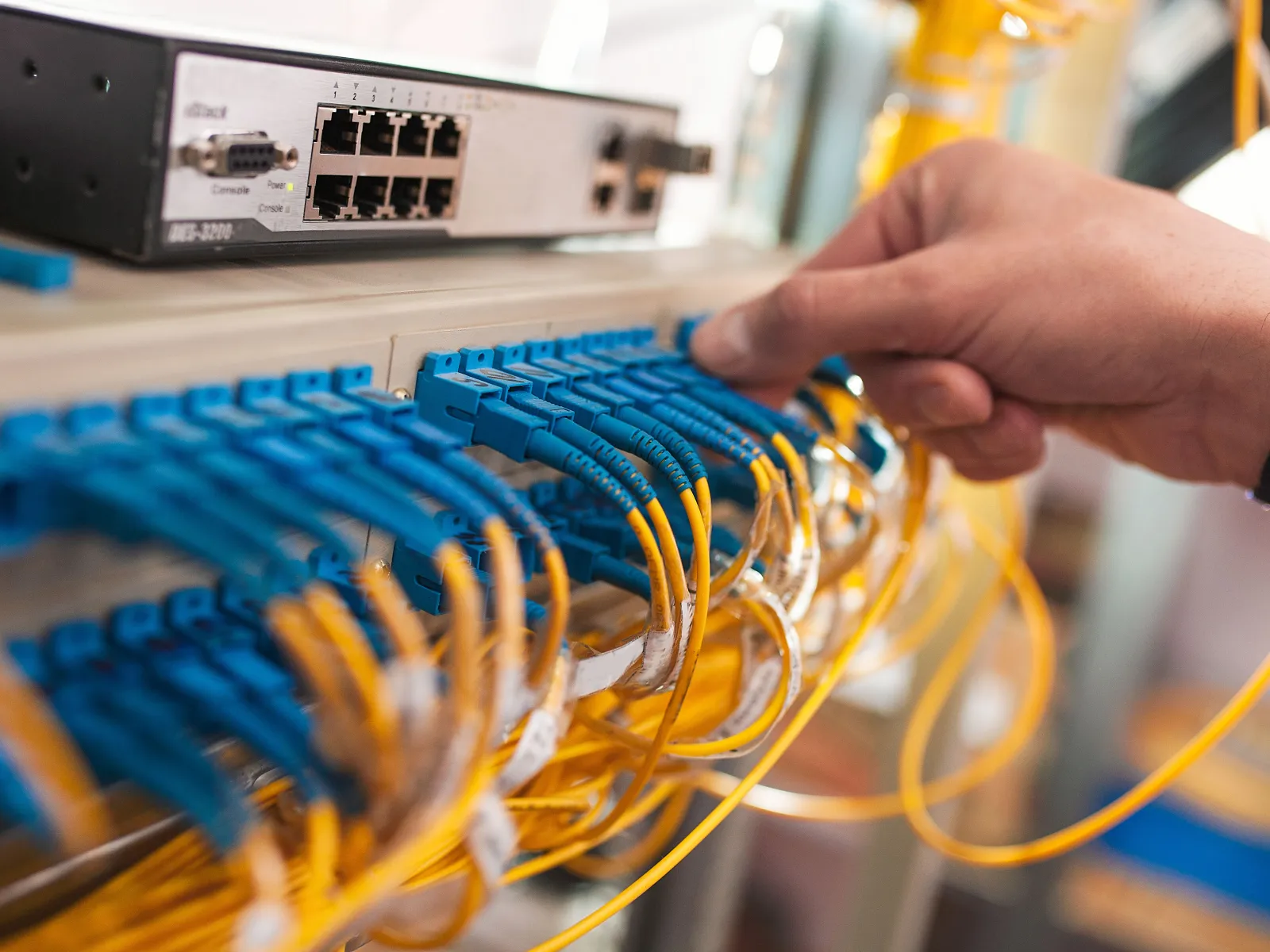 Hand connecting blue fiber optic cables to a network switch with yellow cables in a server room