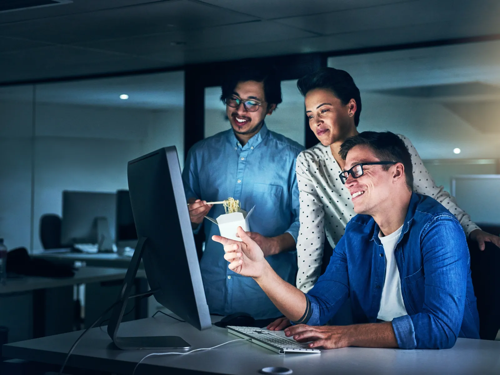 Three coworkers smiling and working together late at night on a computer in a modern office.