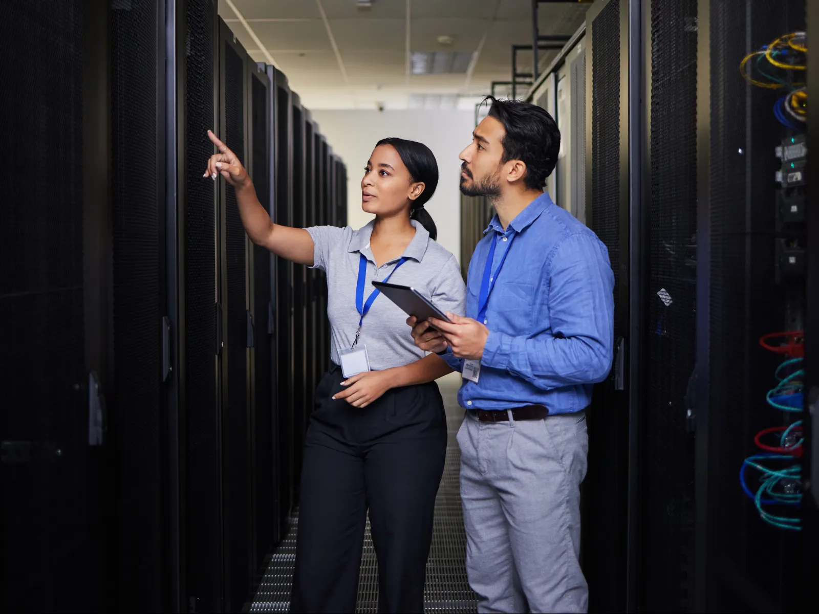 Two IT professionals inspect server racks in a data center, one pointing and the other holding a tablet device.