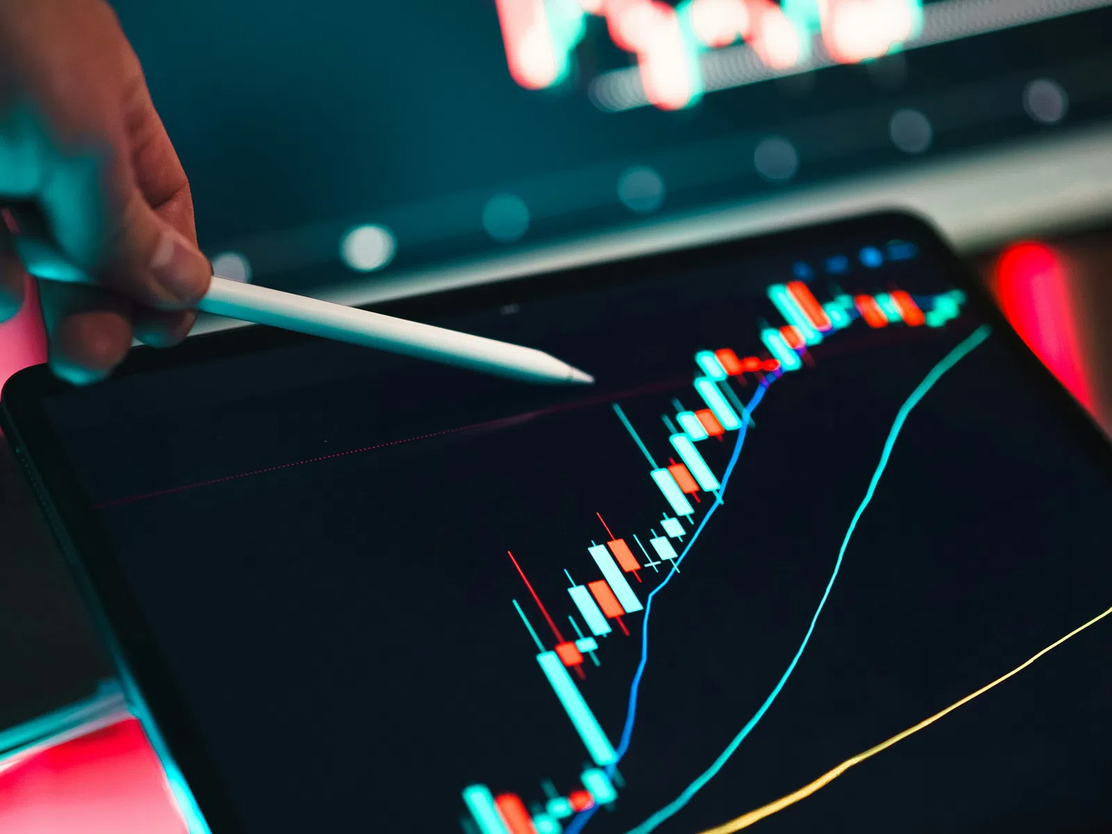 Hand holding stylus pointing at rising financial candlestick chart on tablet with blurred stock graph in background.