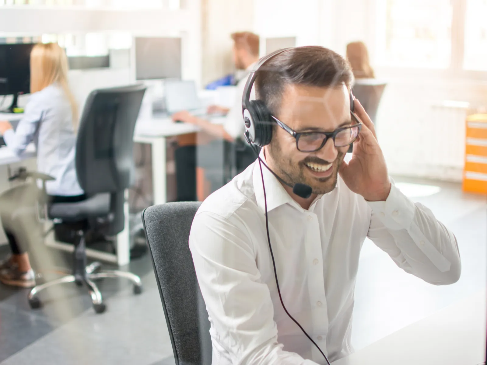 Smiling male customer service agent with headset working at modern office desk with colleagues in background