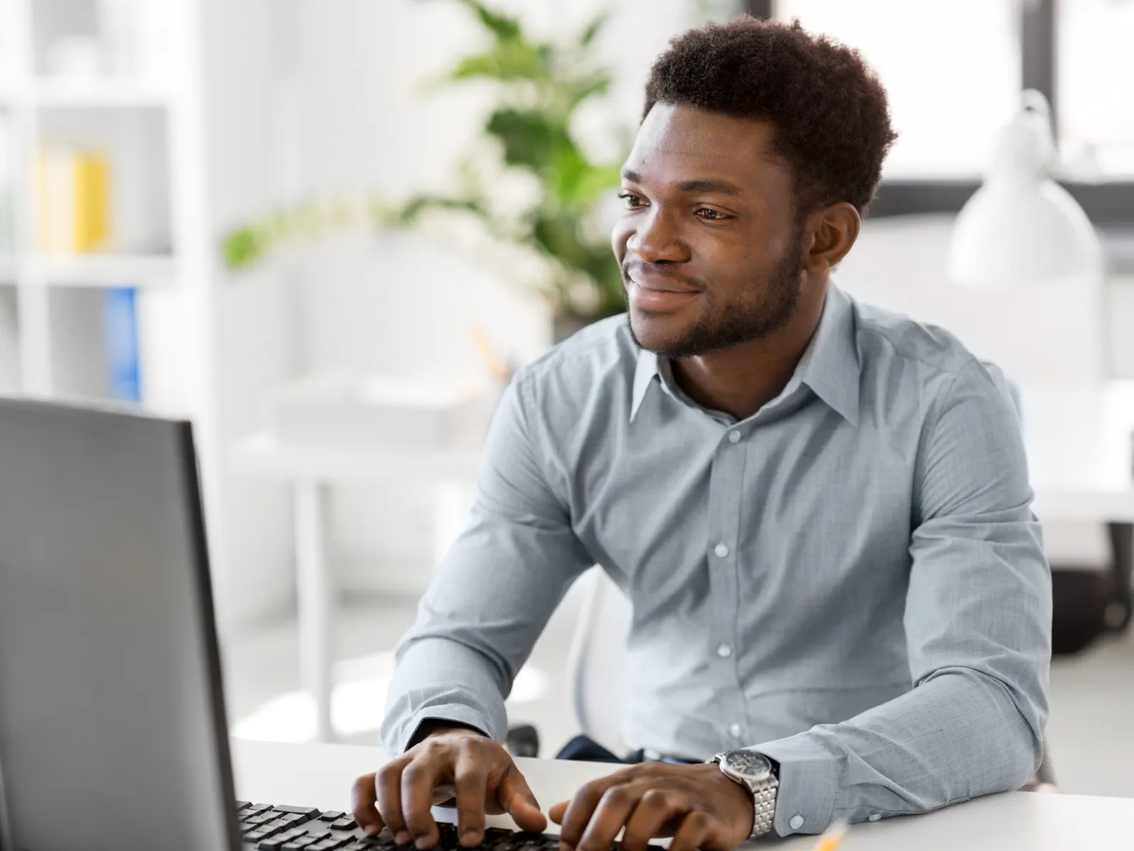 Young man in light blue shirt working at computer in bright modern office with green plant in background