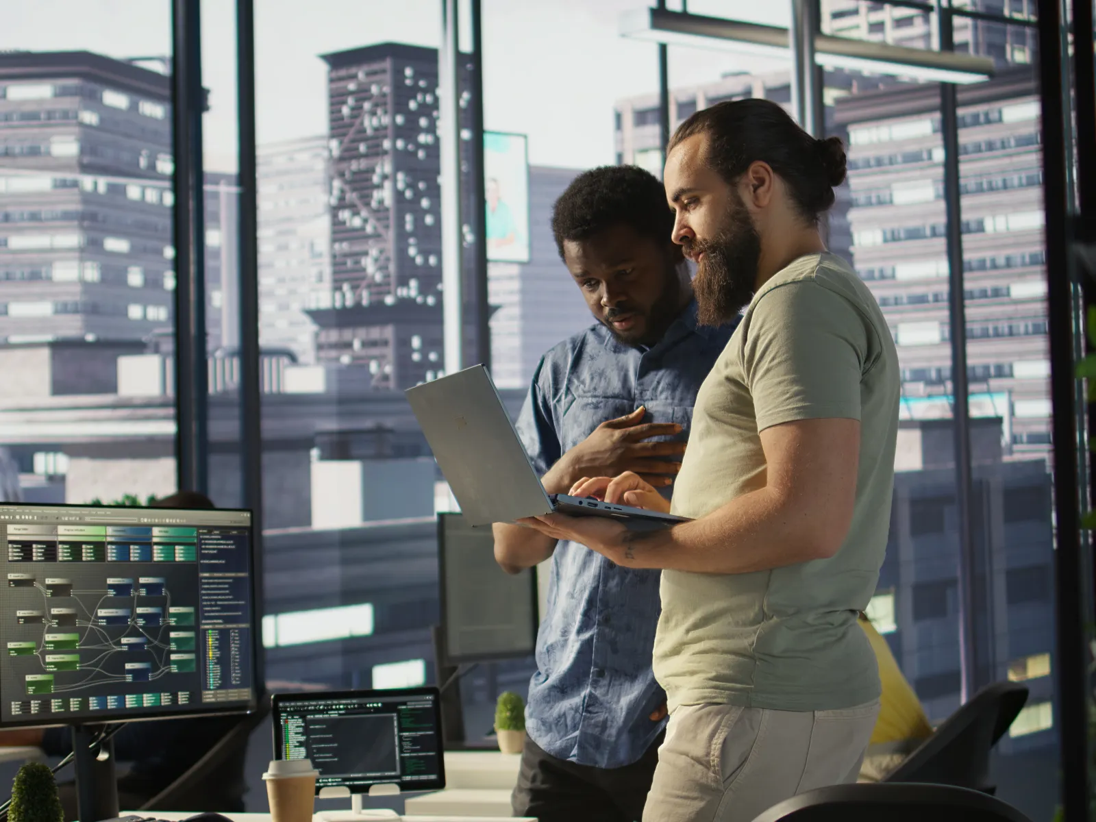 Two developers collaborate in a modern office with city view, using laptop and multiple monitors with coding visuals.