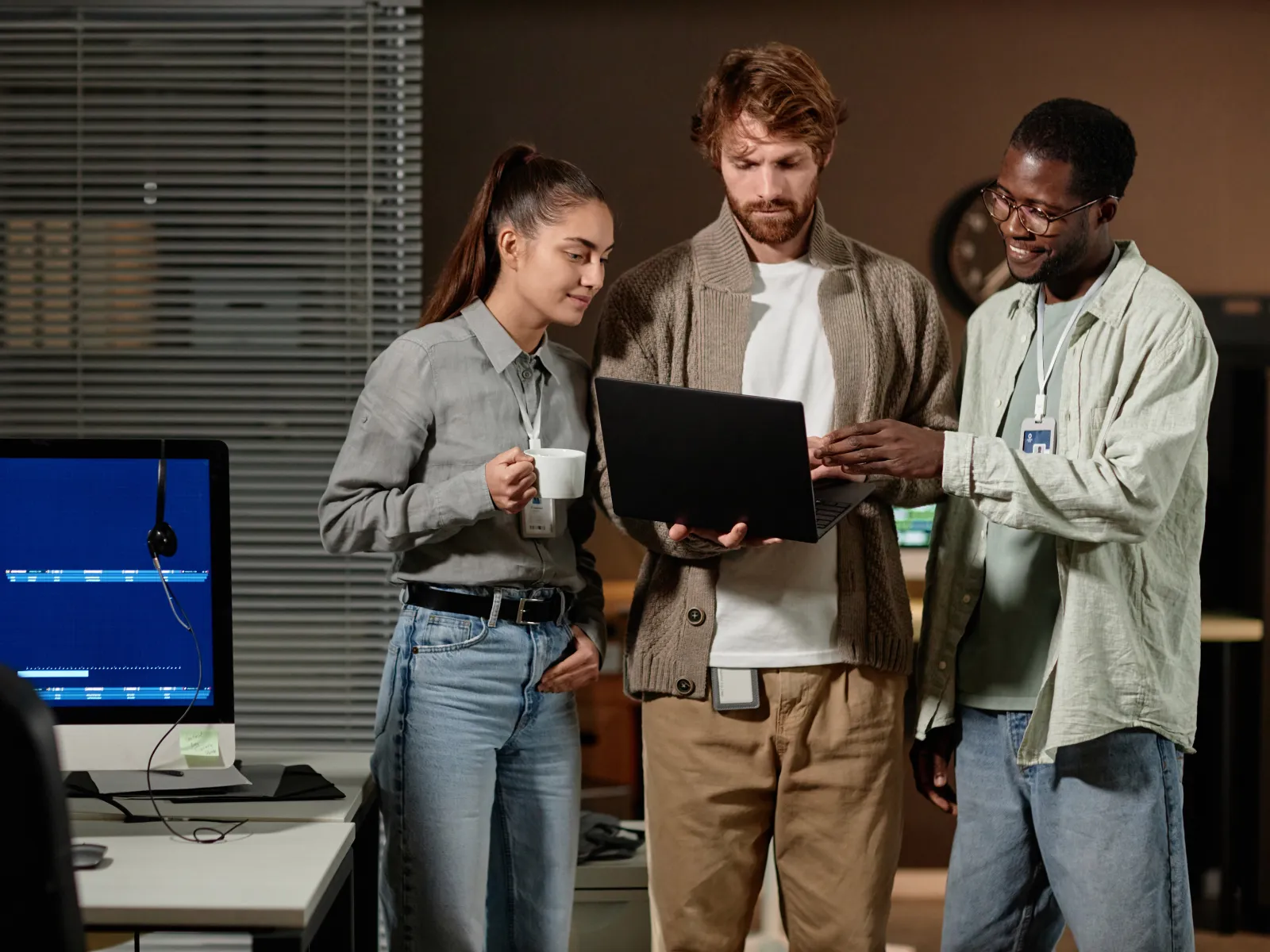 Three coworkers discuss a project standing around a laptop in a modern office with a desktop screen nearby.