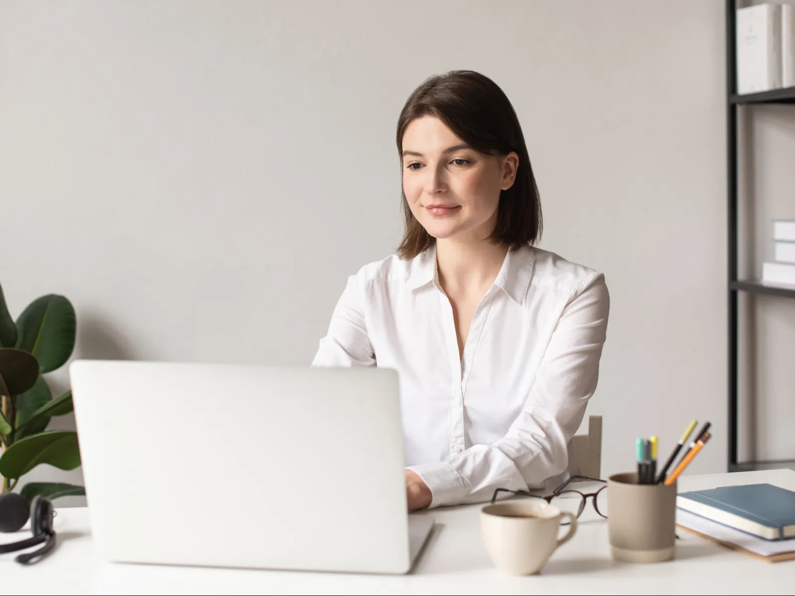 Young woman in white shirt working on a laptop at a clean desk with plant and stationery.