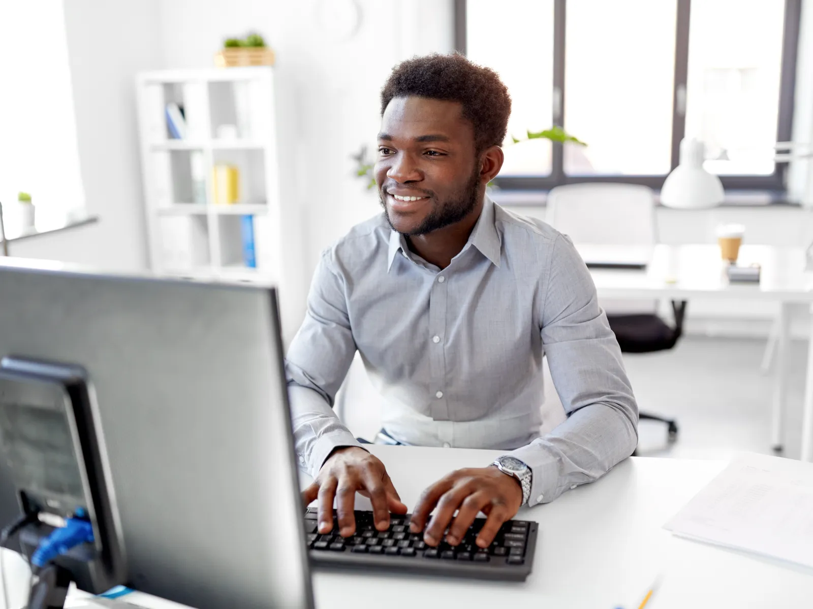 Man smiling while typing on a keyboard in a bright modern office with large windows and white furniture.