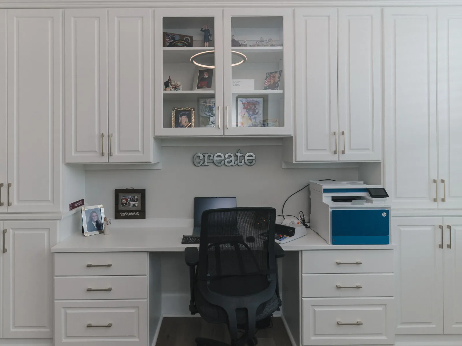 Home office with white cabinets, glass doors, laptop, printer, black executive chair and decorative create sign on the wall