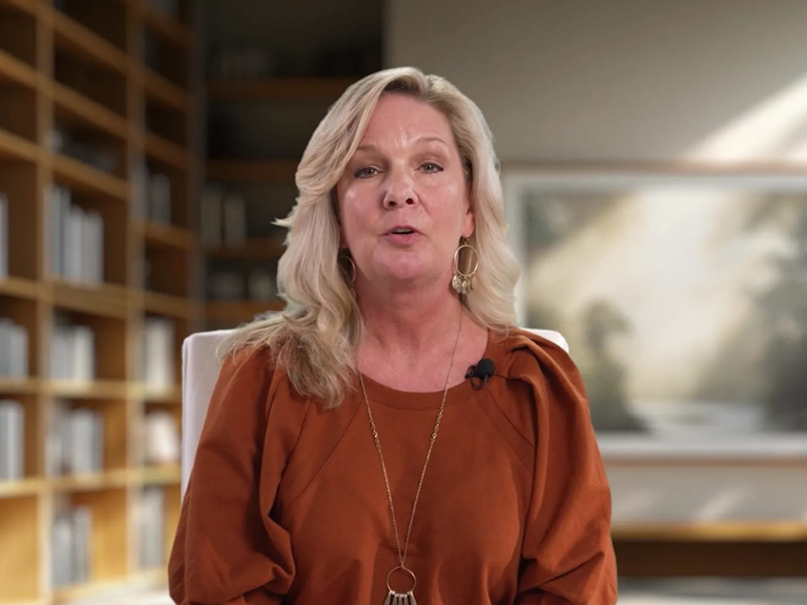 Blonde woman in rust-colored top speaking in a well-lit room with bookshelves and framed art in background