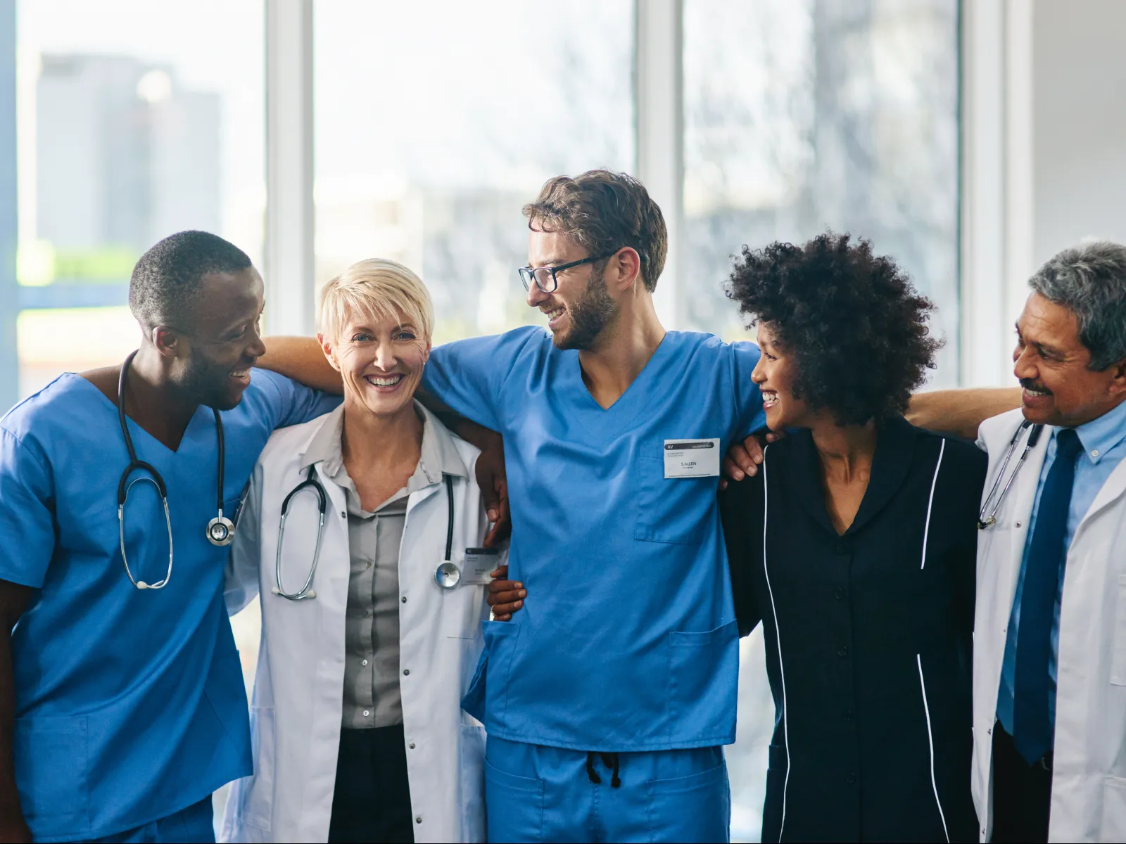 Diverse group of smiling medical professionals standing together with arms around each other in hospital hallway.