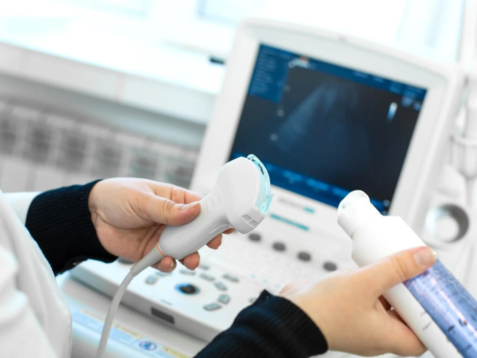 Close-up of hands holding an ultrasound probe and gel bottle in front of an ultrasound machine screen.