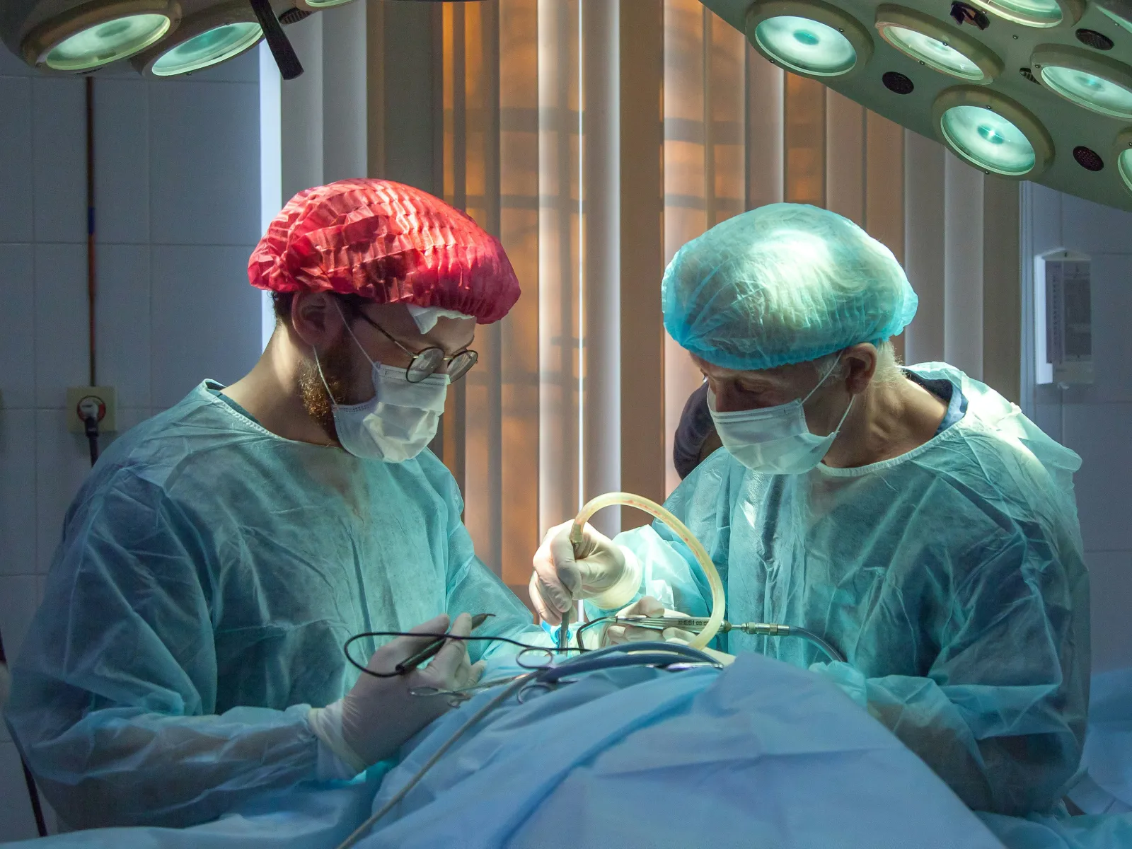 Two surgeons wearing protective gear perform surgery under bright operating room lights.