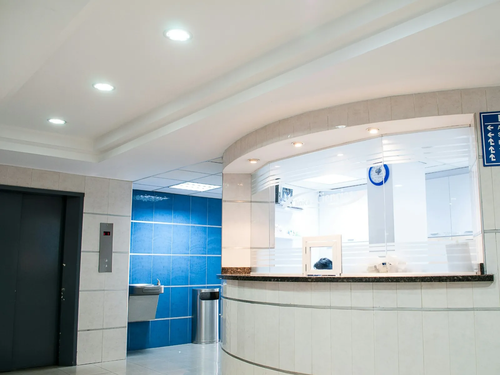 Bright hospital reception area with white walls, a counter window, elevator, and directional signage in Spanish.