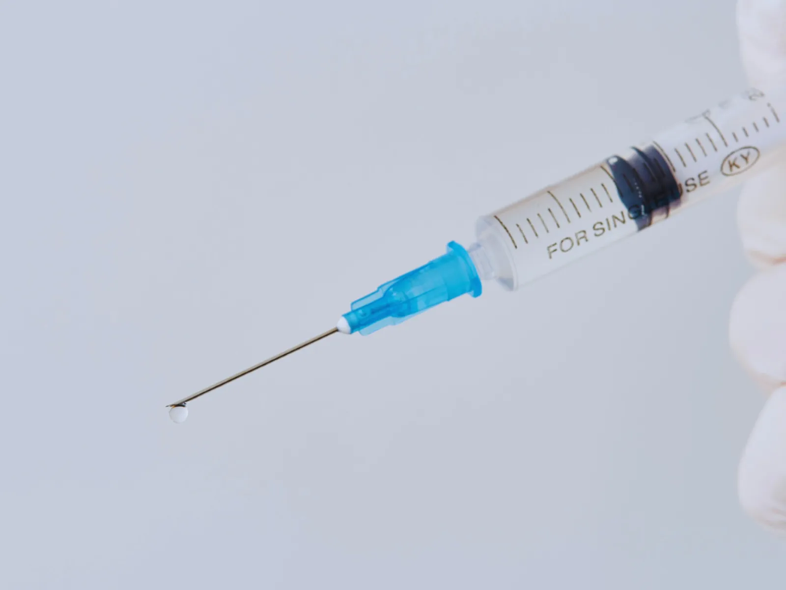 Close-up of a syringe with a drop at the needle tip held by a gloved hand against a light background