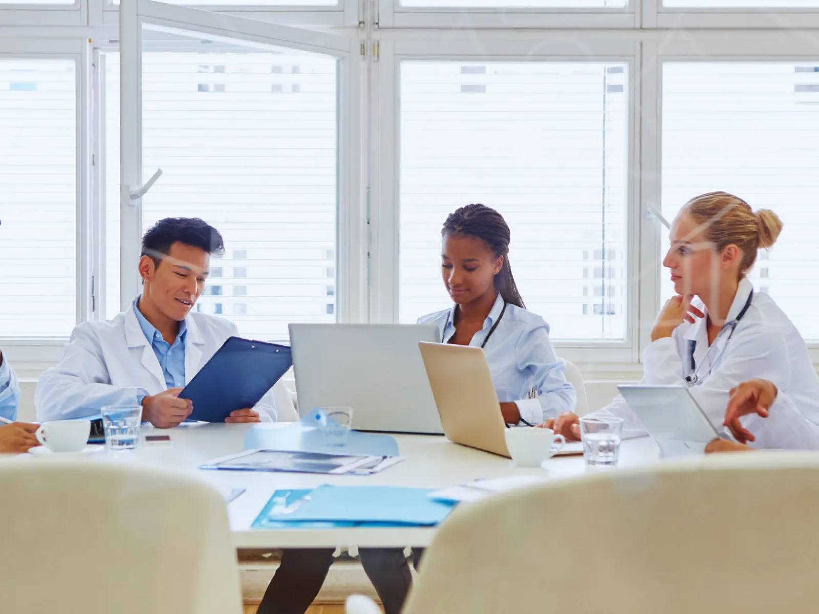 Diverse medical team in white coats collaborating with laptops and documents in bright conference room