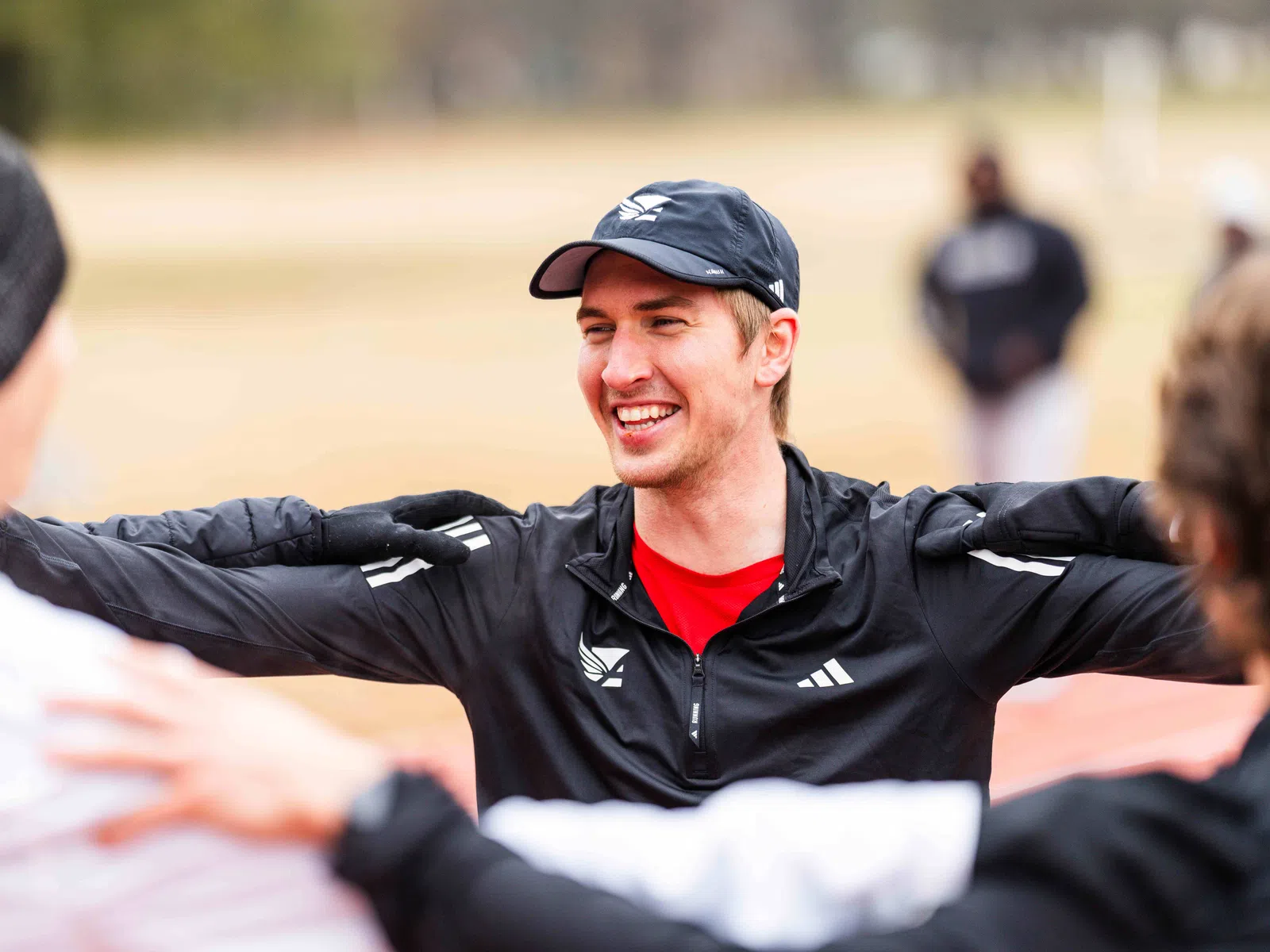 Smiling young man in black jacket and cap stretching arms outdoors with friends during exercise session