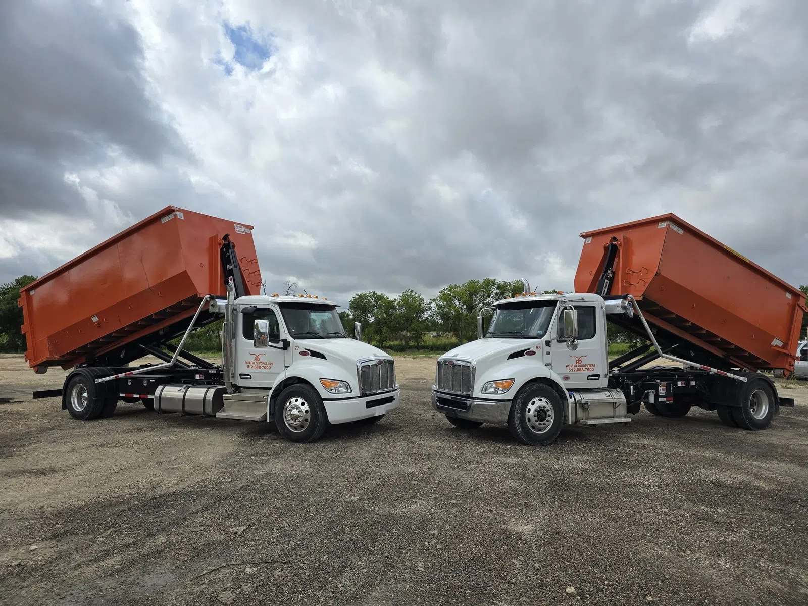Two white dump trucks with raised orange containers parked on a gravel lot under a cloudy sky.