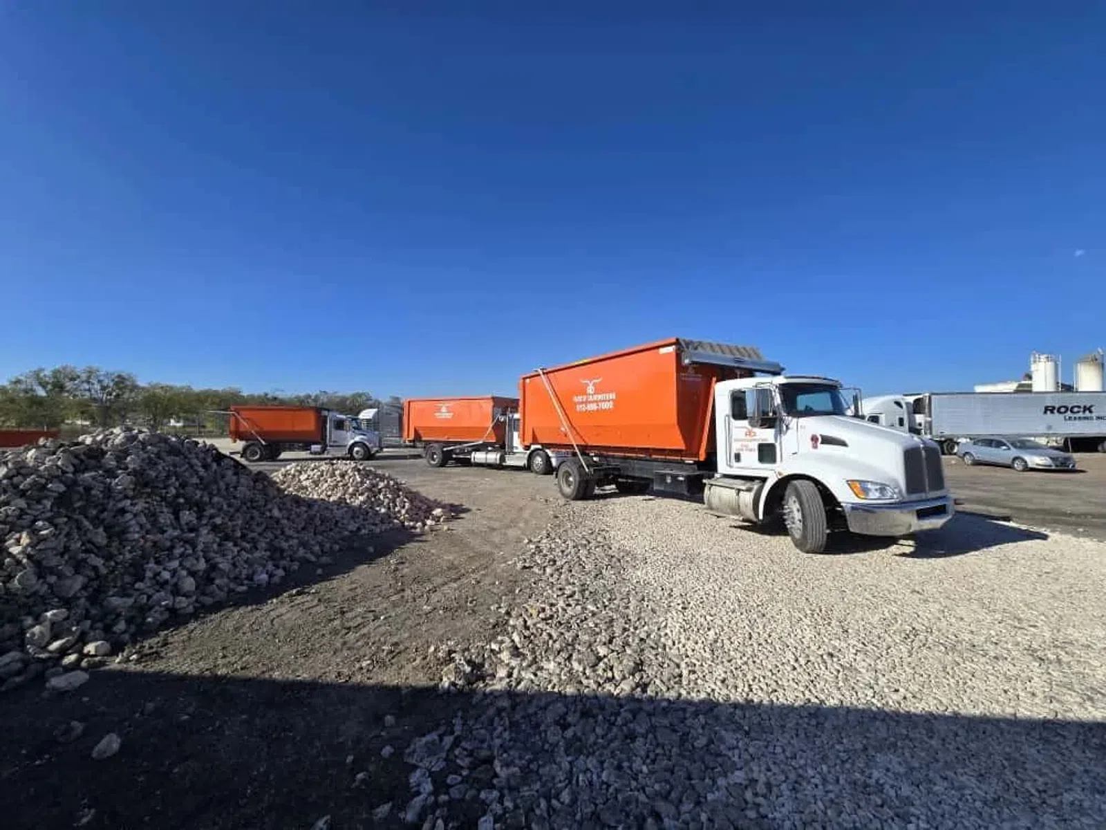 White trucks with bright orange containers parked on gravel near stone piles under clear blue sky.