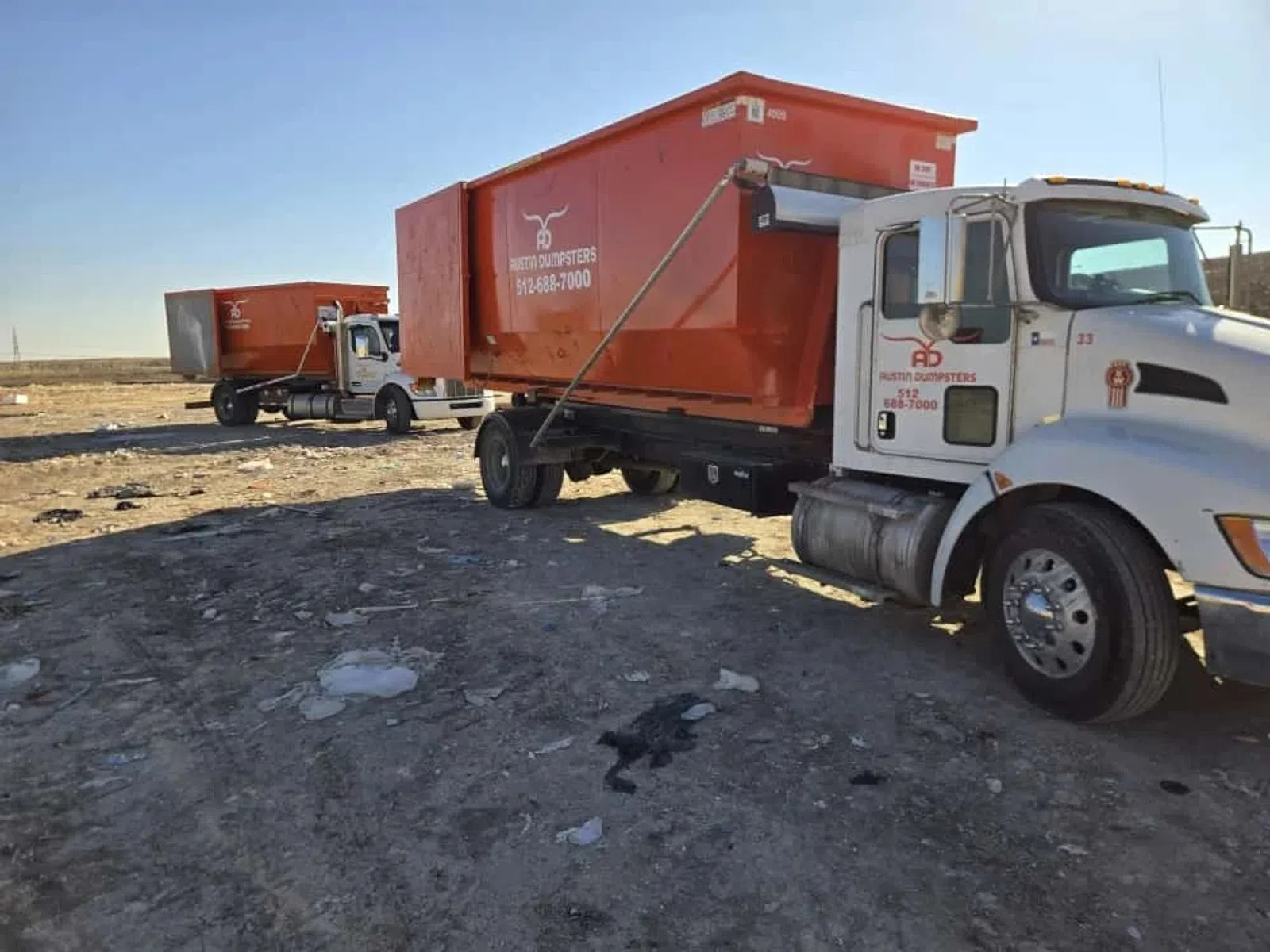 Two white trucks carrying large red dumpster containers on a dirt lot under a clear blue sky