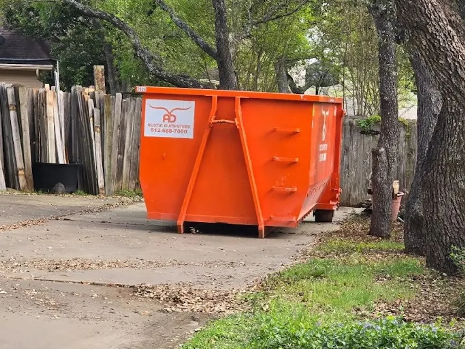 Bright orange Austin Dumpsters container on a driveway near trees and wooden fence in a residential area.