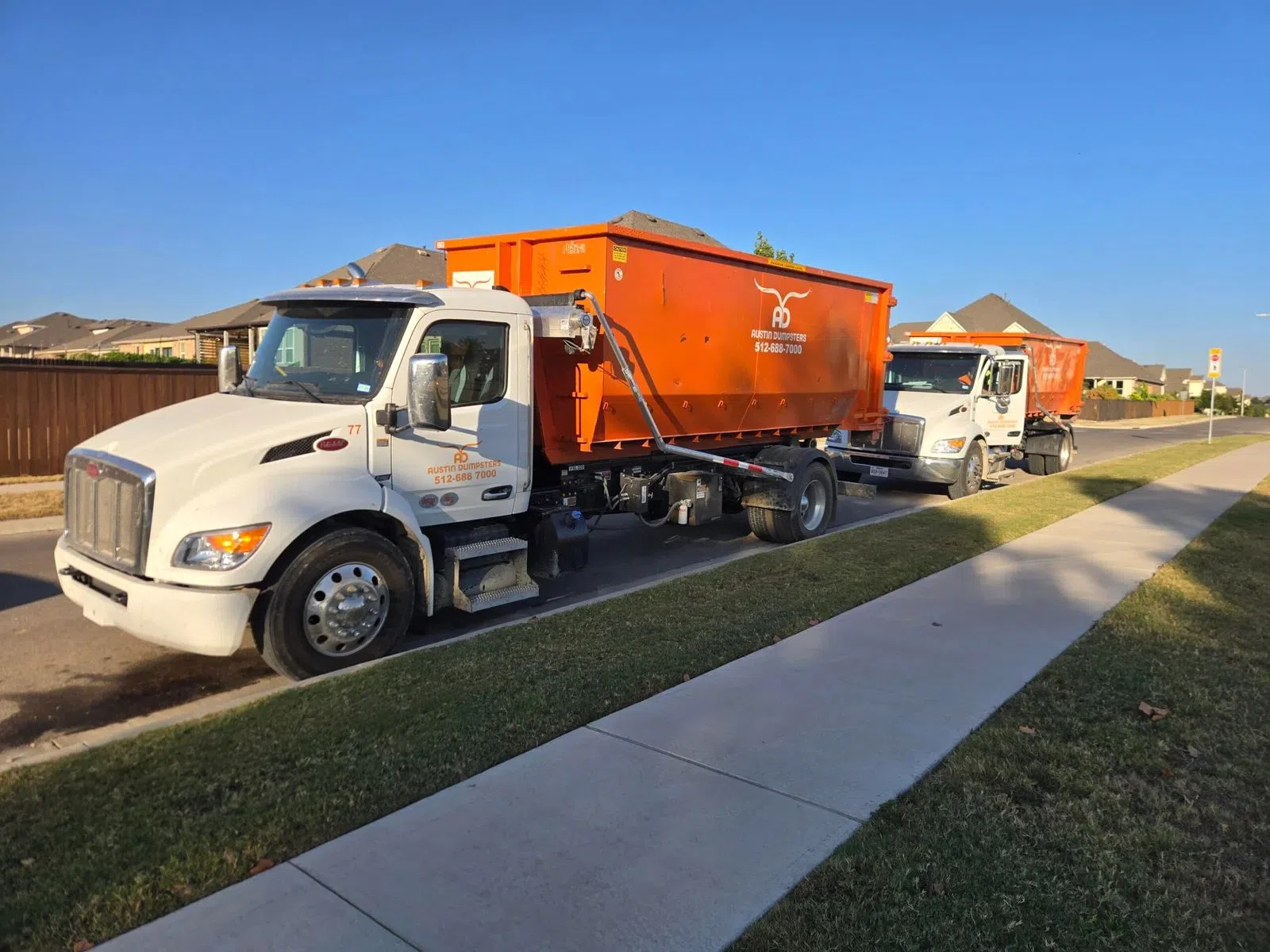 Two white trucks with large orange dumpsters parked on a residential street next to a sidewalk under clear blue sky.