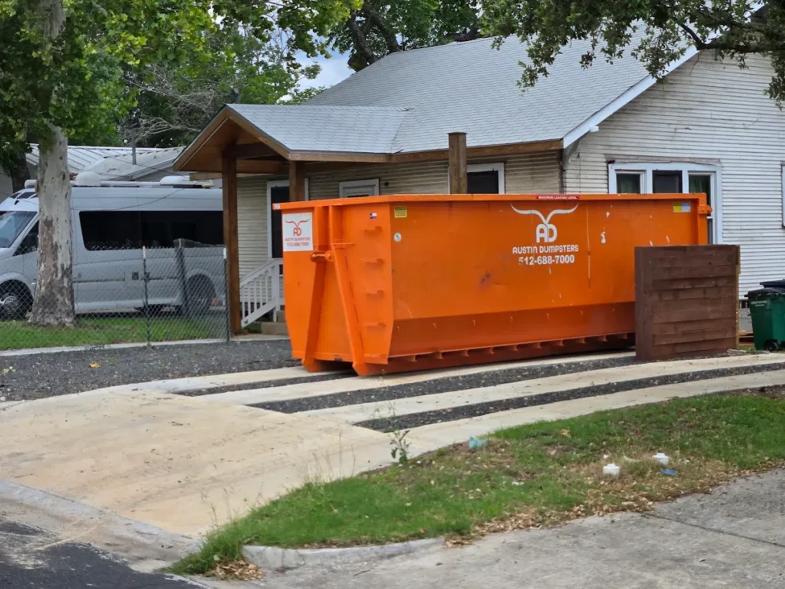 Large orange dumpster from Austin Dumpsters placed on a concrete pad in front of a white house with trees.