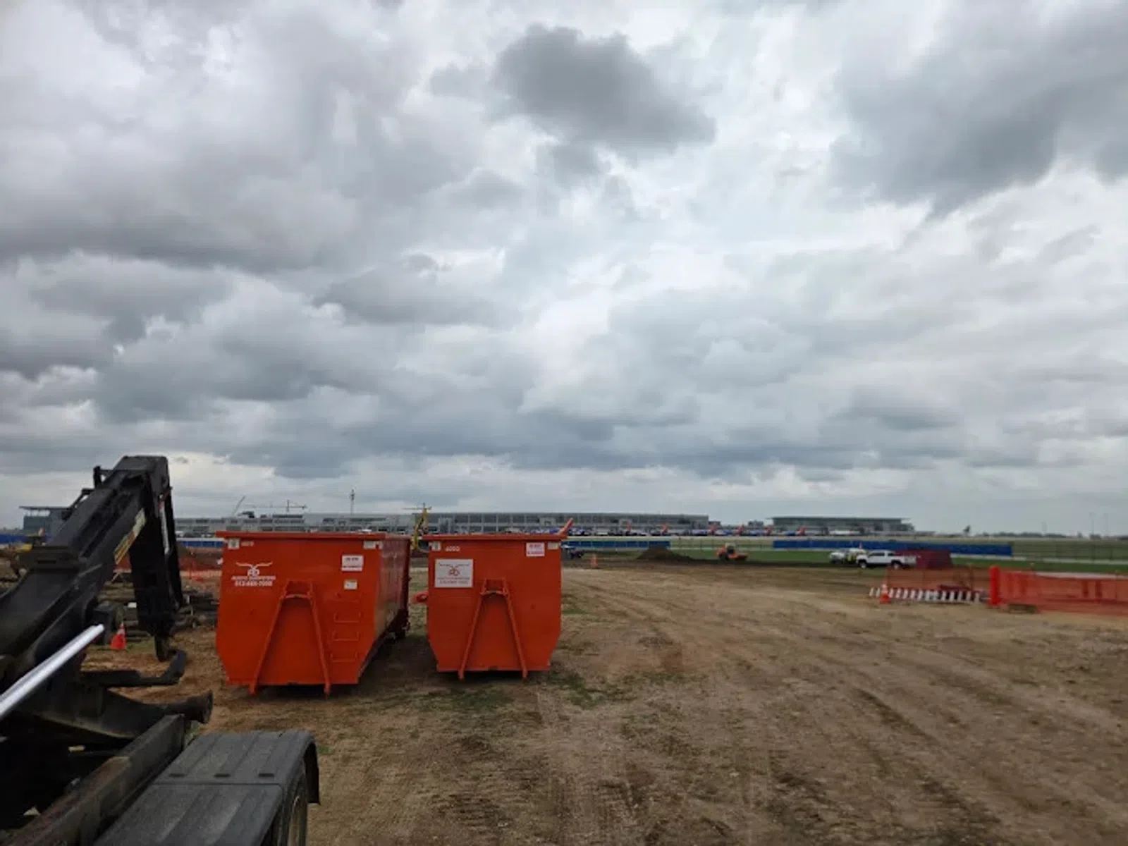 Construction site with two orange dumpsters under cloudy sky on a dirt field near industrial buildings.
