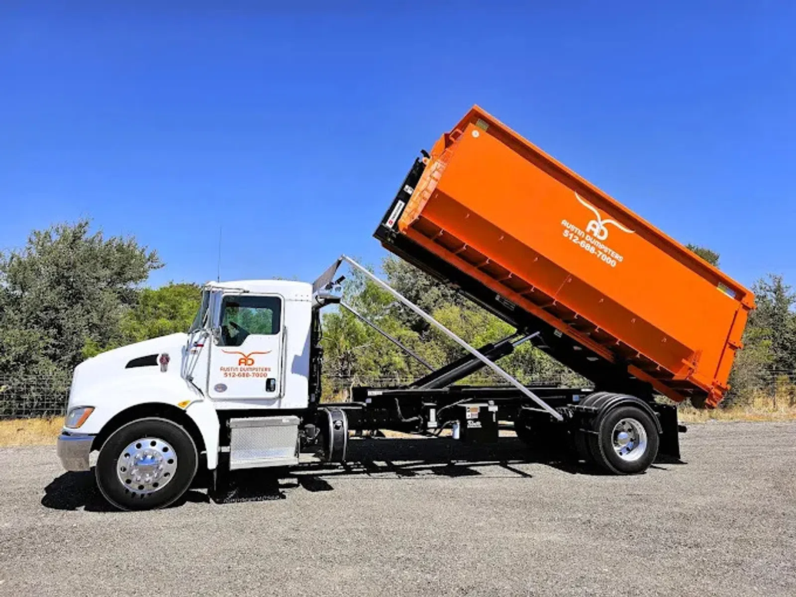 White truck with tilted orange dumpster container on a gravel lot under clear blue sky.