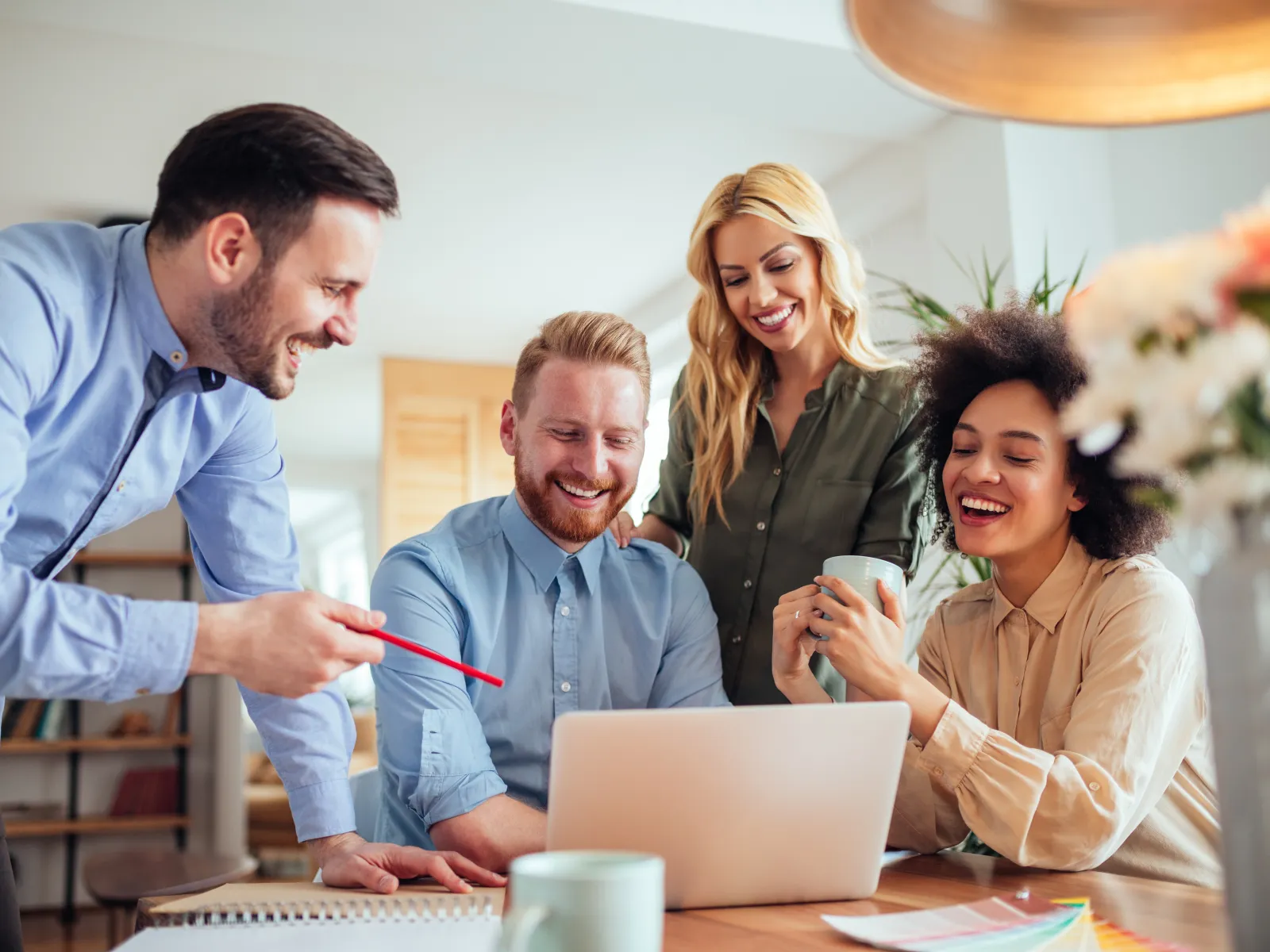 Four diverse professionals smiling and collaborating around a laptop in a bright modern office.