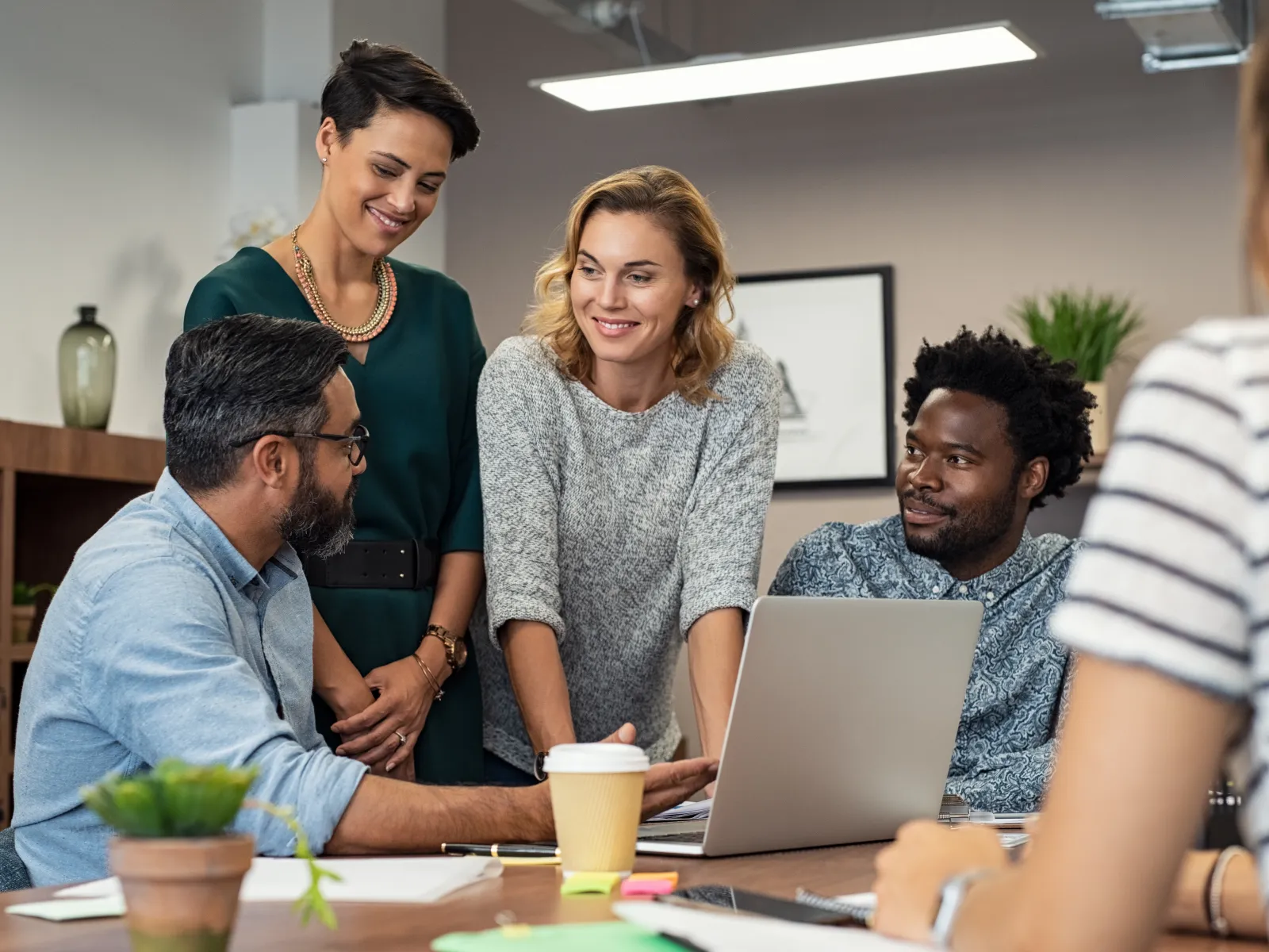 Diverse group collaborating around a laptop in a modern office with casual and professional attire engaging in discussion.