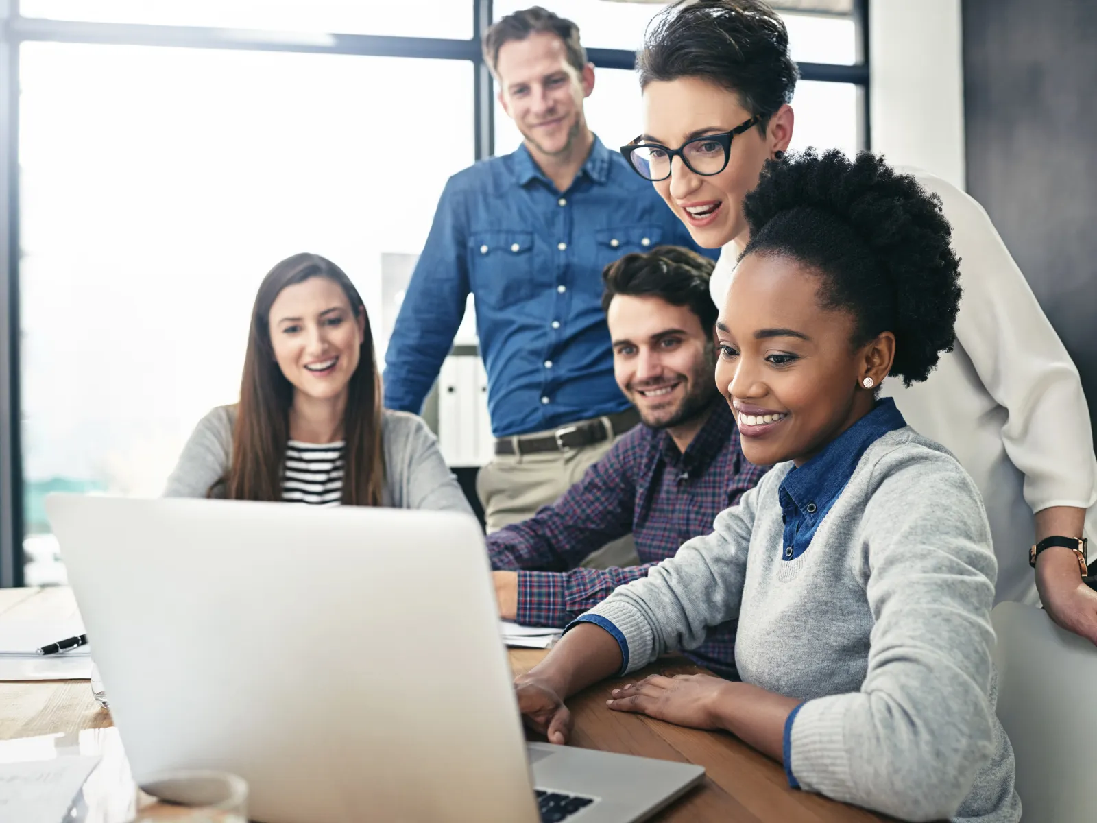 Diverse group of five coworkers collaborating and smiling while looking at a laptop screen in bright office.
