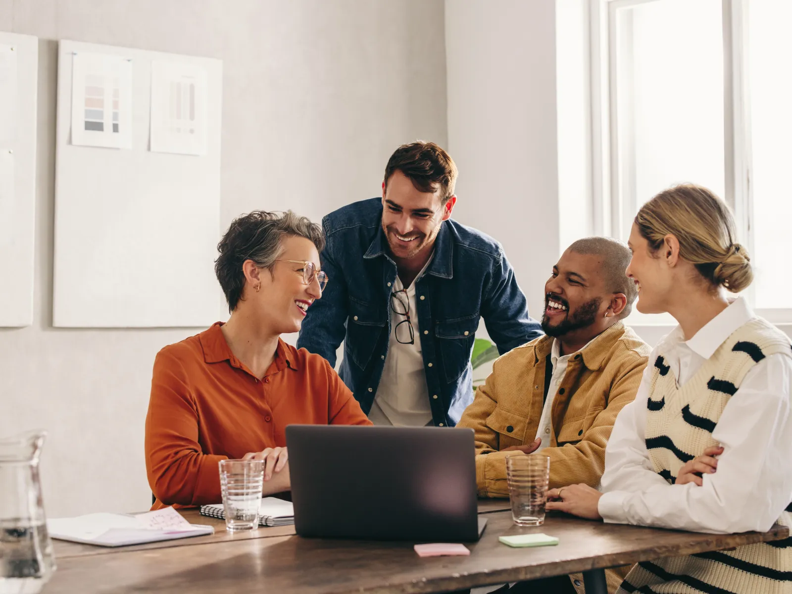 Four diverse colleagues smiling and collaborating around a laptop in a bright modern office meeting room.