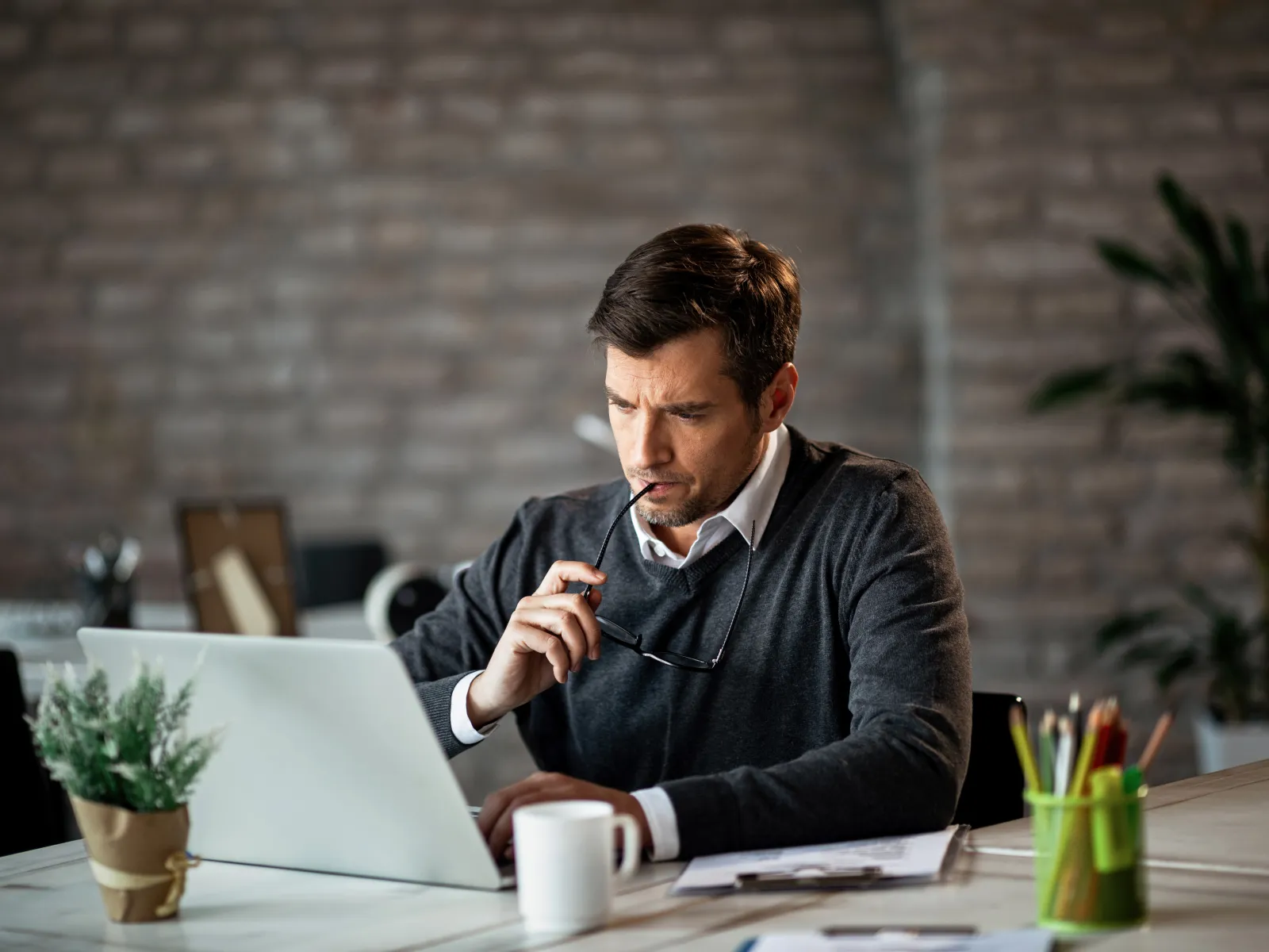 Man in sweater working on laptop at desk with coffee cup and plant in cozy office with brick wall.